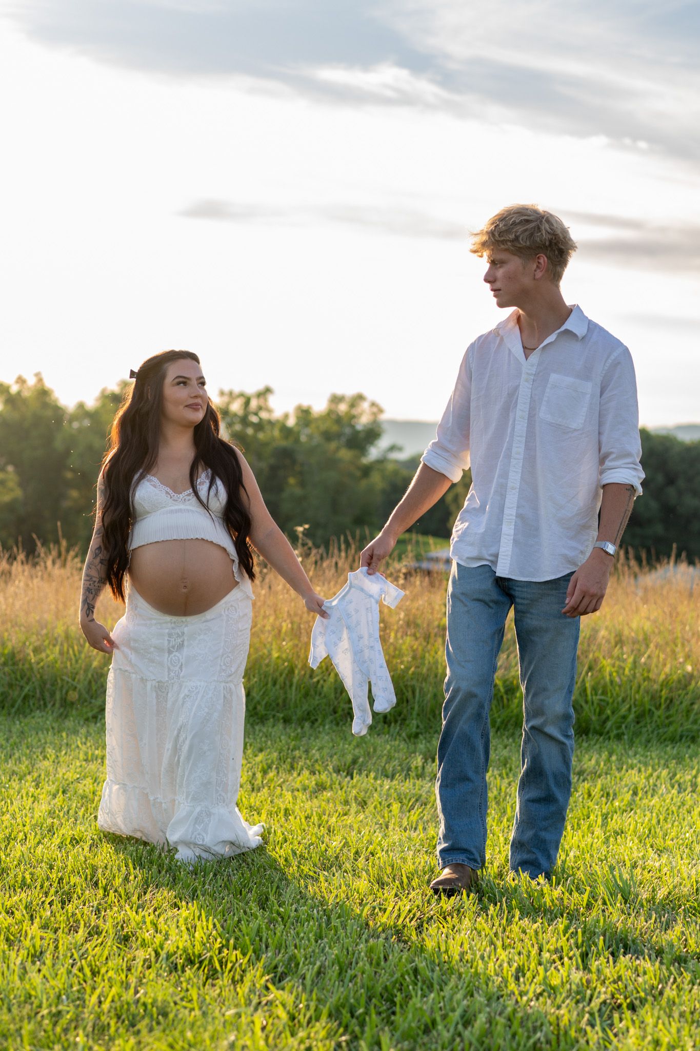Expecting couple holding baby clothes while walking hand in hand through green field at sunset.
