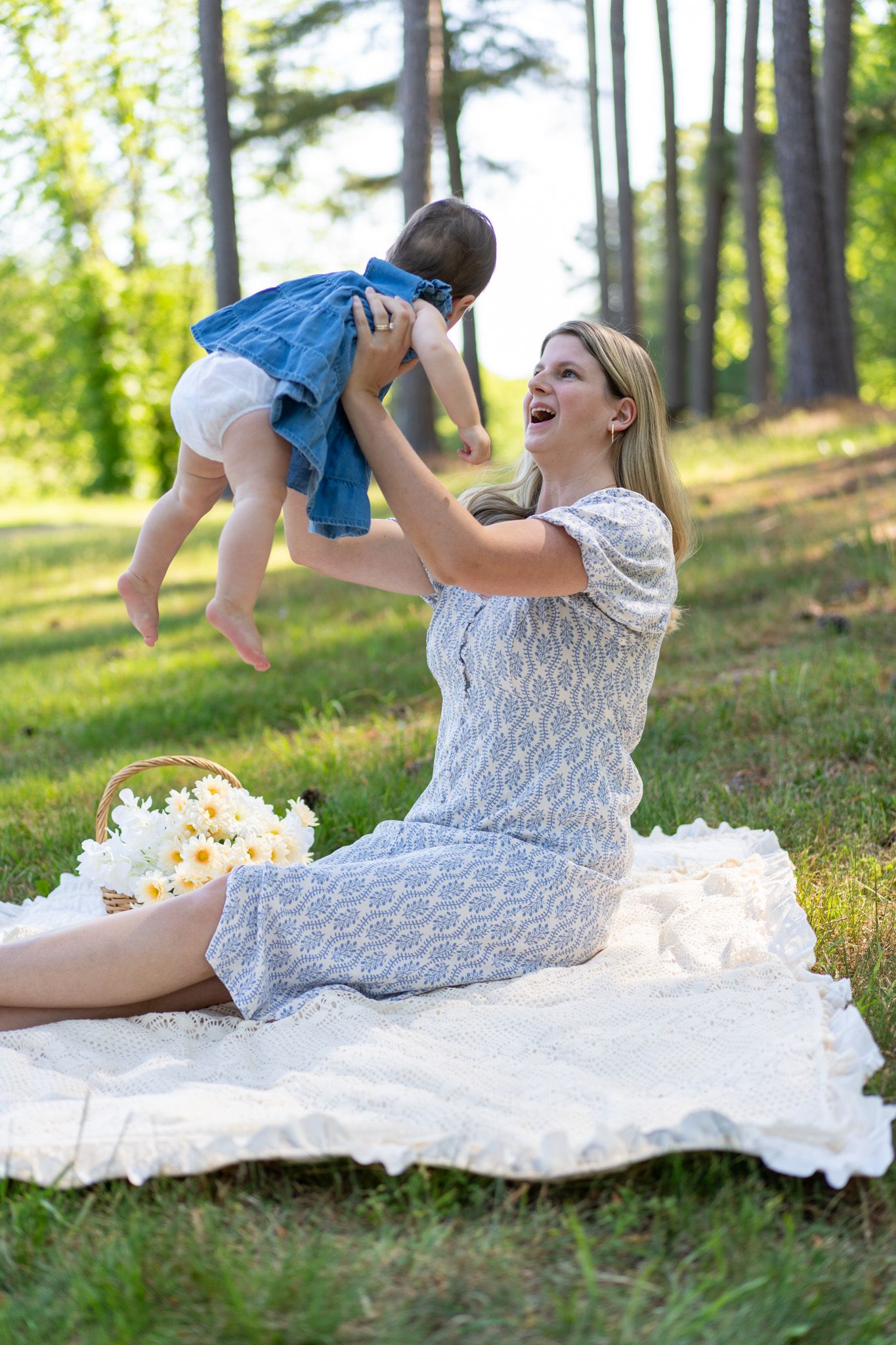 Laughing blonde mother in white lace dress lifts baby in denim outdoors on blanket in sunny park.