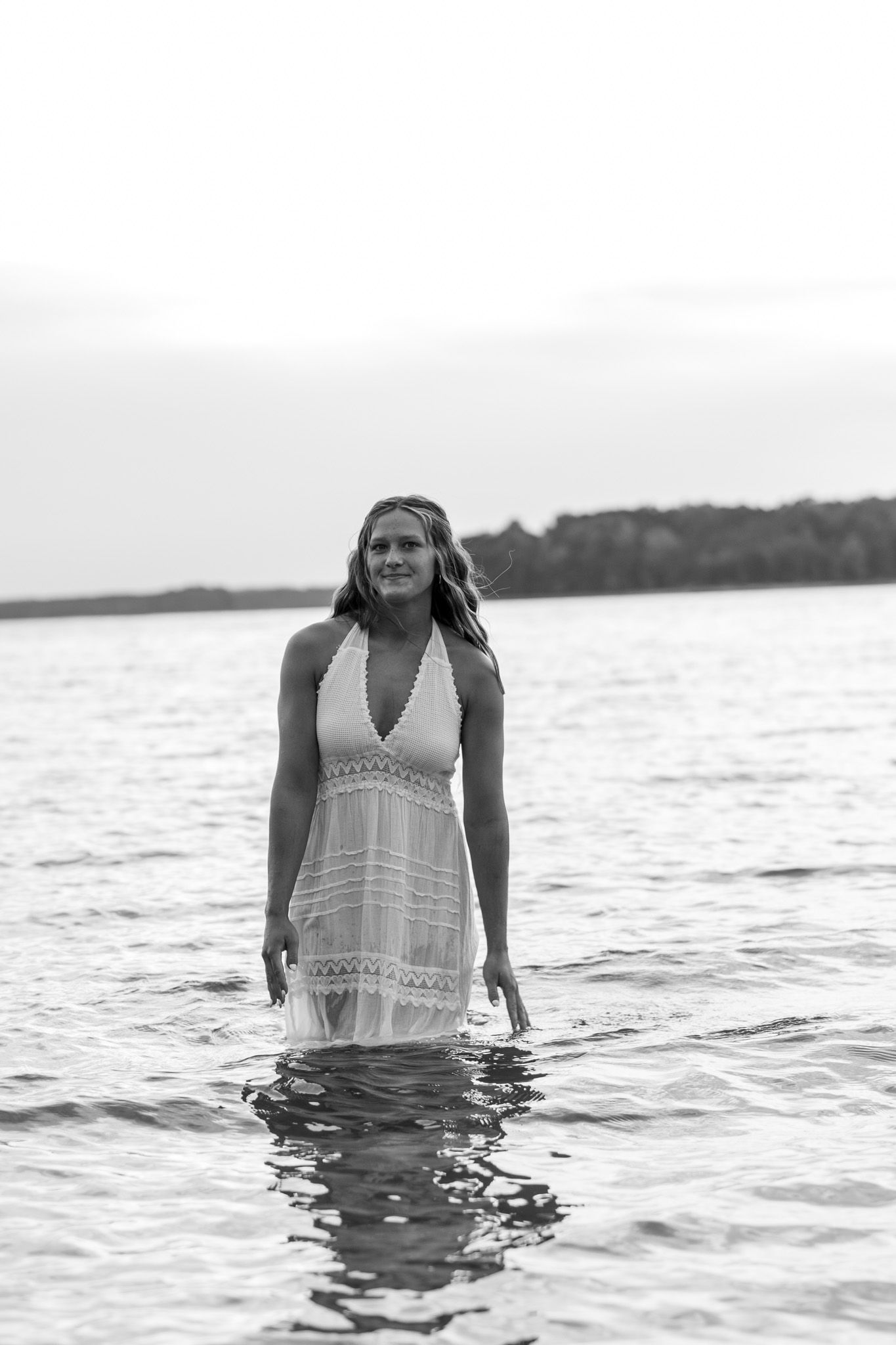 Black and white photo of a young woman in a white halter dress standing in calm lake water at dusk.