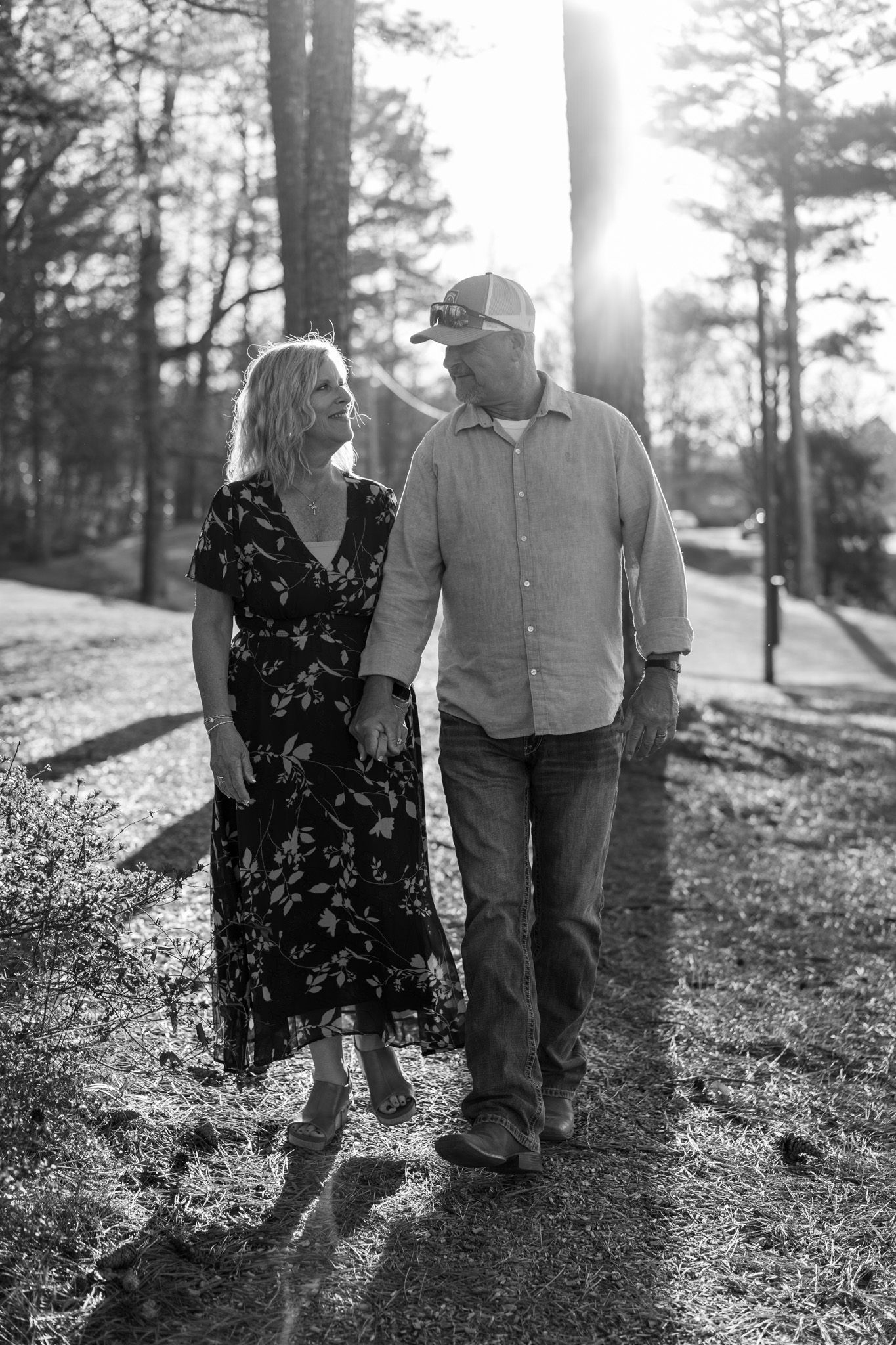 Black and white photo of a smiling couple walking together through a sunlit wooded park.