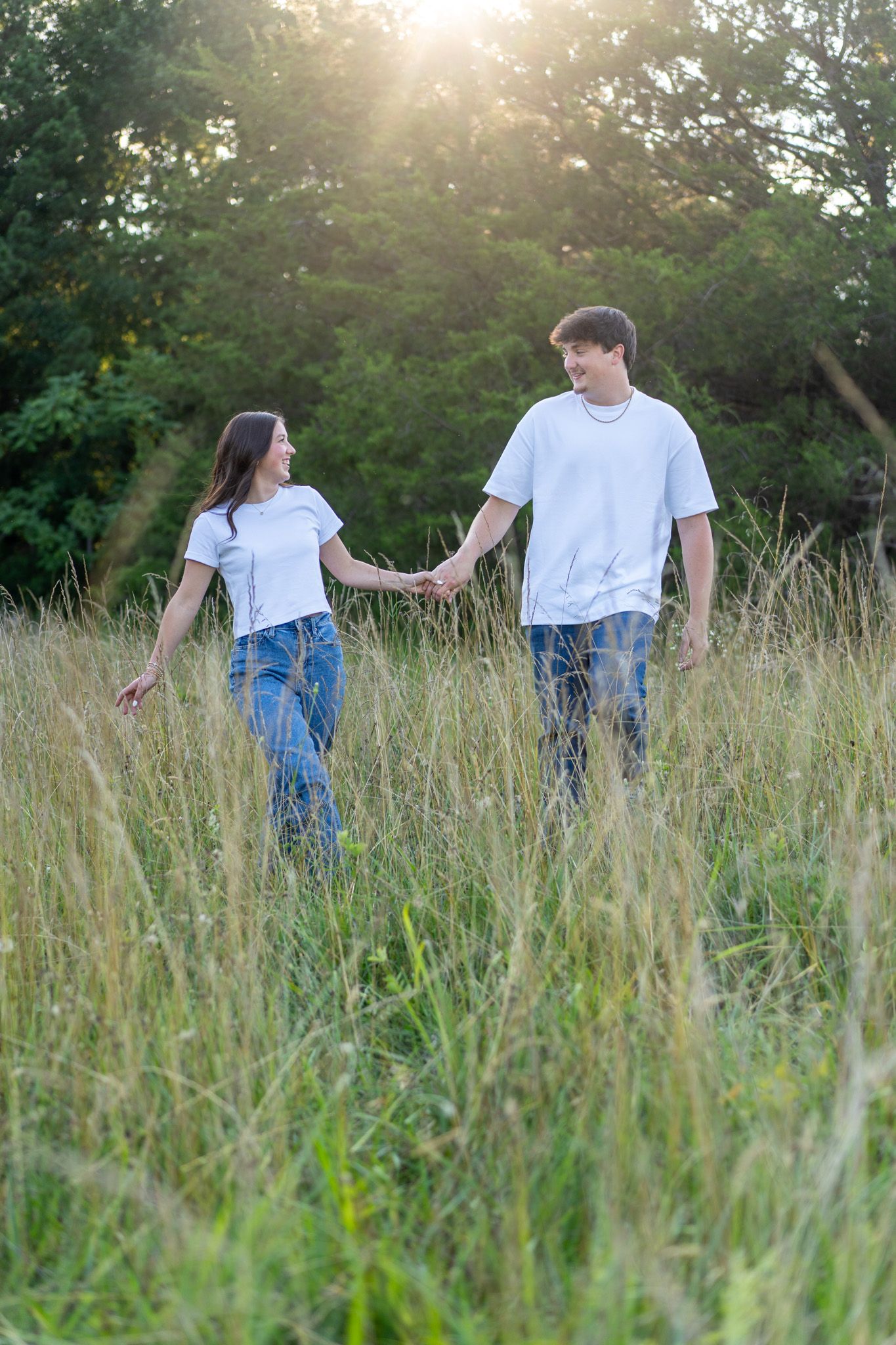 A young couple in matching white tees and jeans holds hands while walking through a sunlit grassy field.