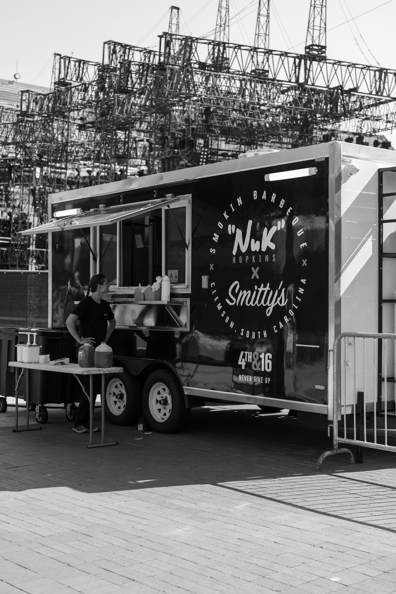 Black and white photo of Smitty's BBQ food truck trailer with worker preparing food, industrial towers in background.