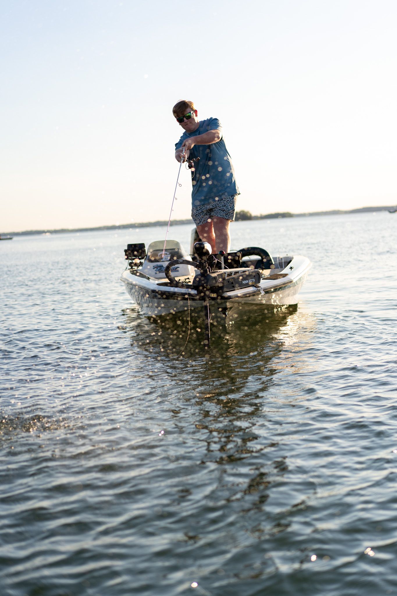 A man stands on a small fishing boat reeling in a catch on calm open water under a clear sky.