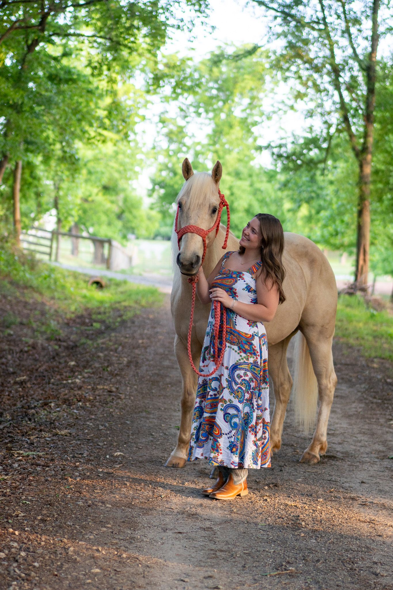 Young woman in floral dress and cowboy boots walking with a palomino horse on a dirt path.