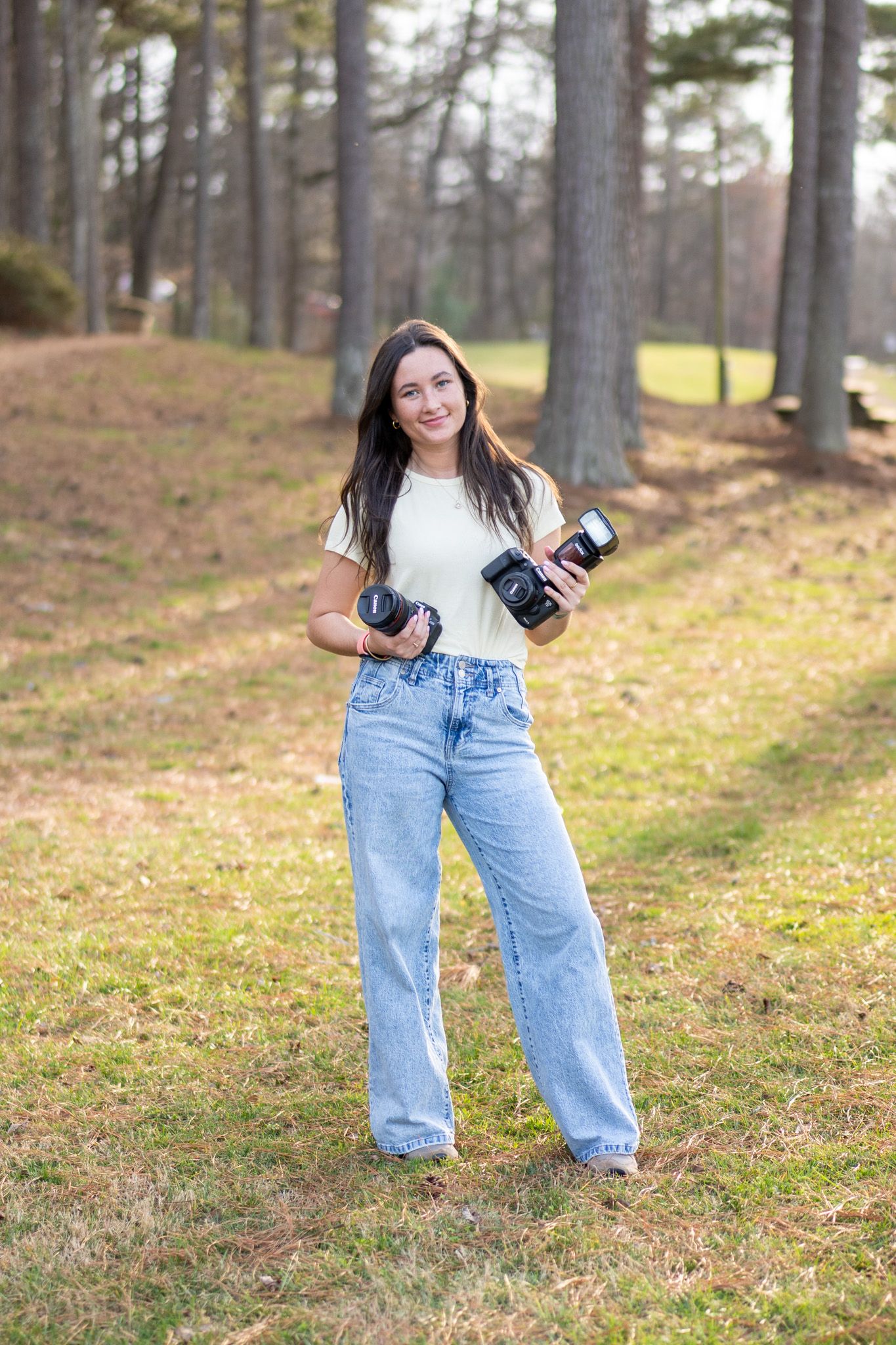 Young woman holding cameras in a wooded park, wearing white tee and wide-leg jeans.