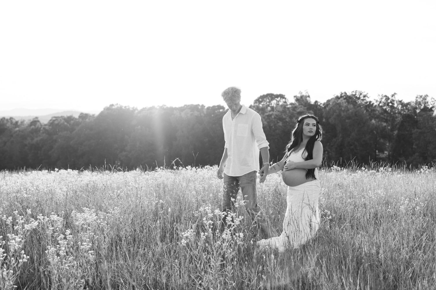 Black and white maternity photo of expecting couple walking through wildflower field in bright sunlight.
