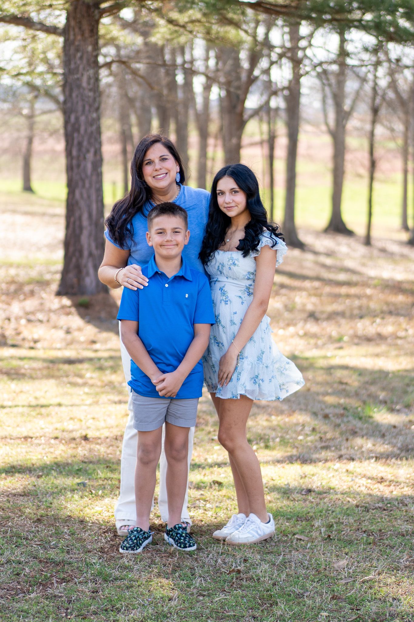 A mother and her two children smile together outdoors, dressed in coordinating blue and white outfits among trees.