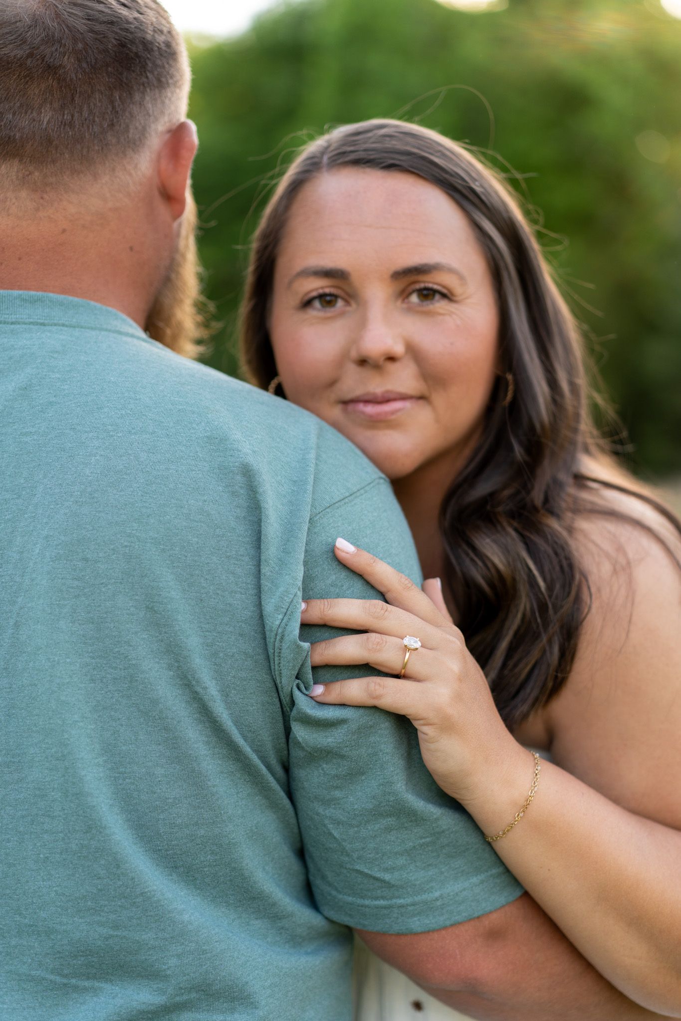 Brunette woman hugging mans shoulder showing engagement ring outdoors.