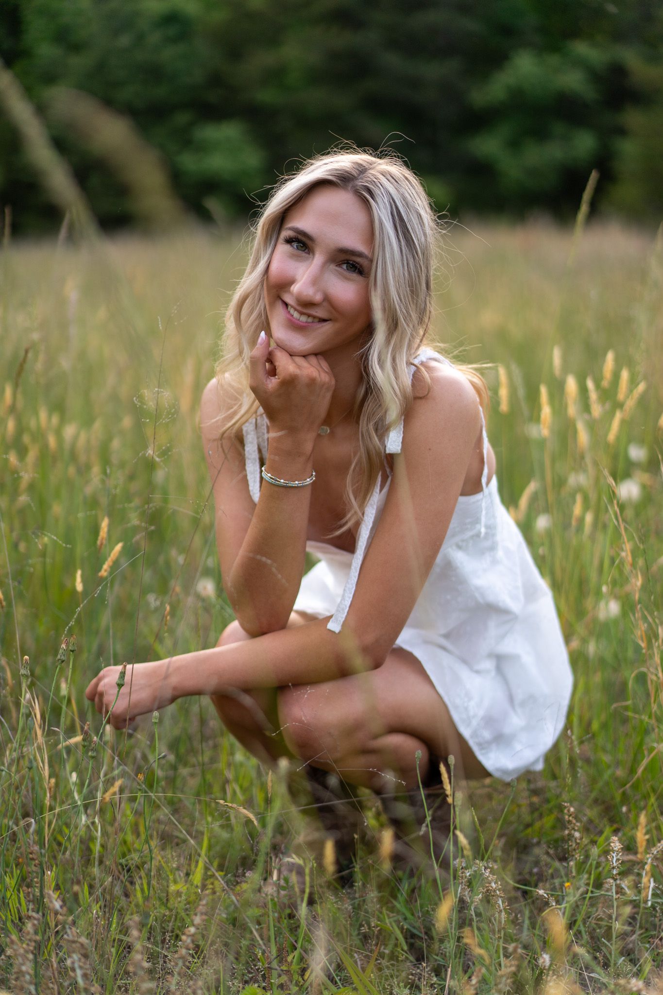 Smiling blonde woman in white dress crouching in tall grass field with green trees in background.