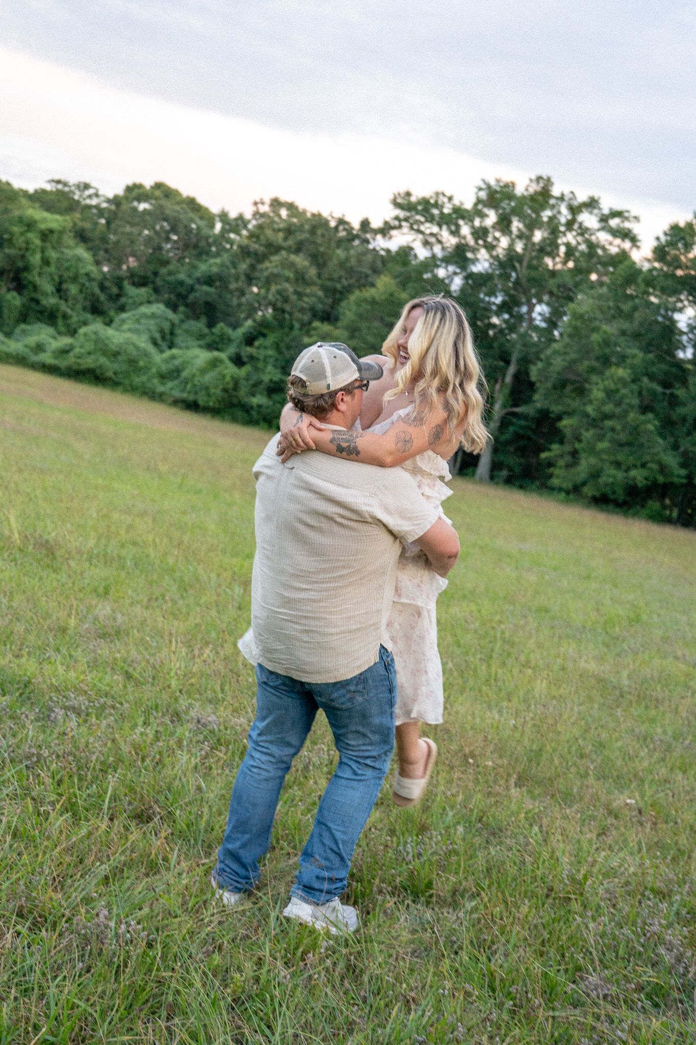 Man in cap lifting and spinning laughing blonde woman in white dress in green meadow at dusk.