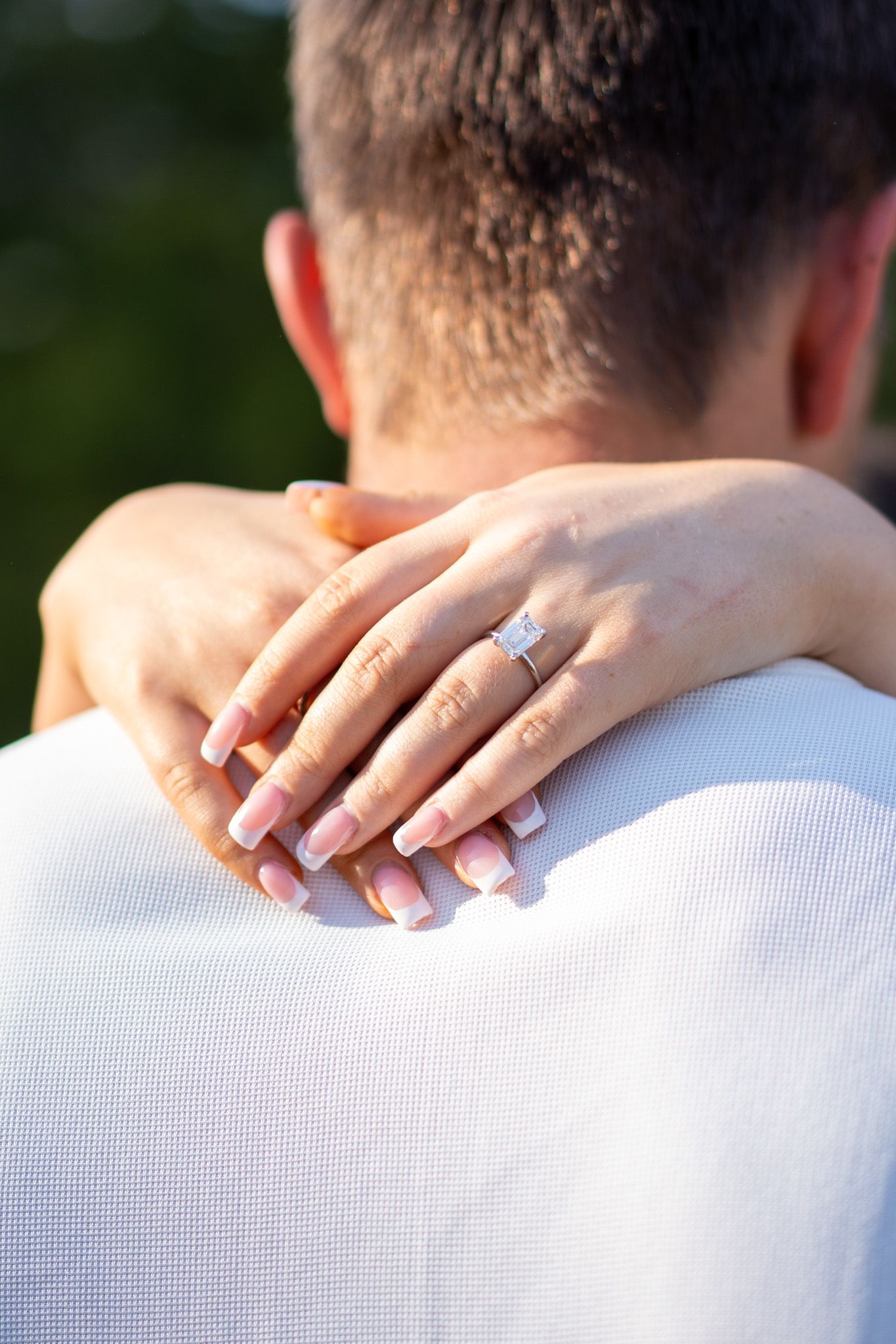 Close-up of woman's hand with diamond engagement ring resting on man's shoulder in white shirt.