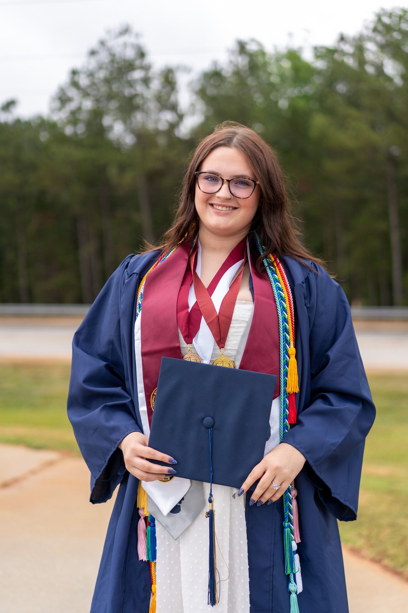 Smiling graduate in navy cap and gown with honor cords and medals holding diploma outdoors.