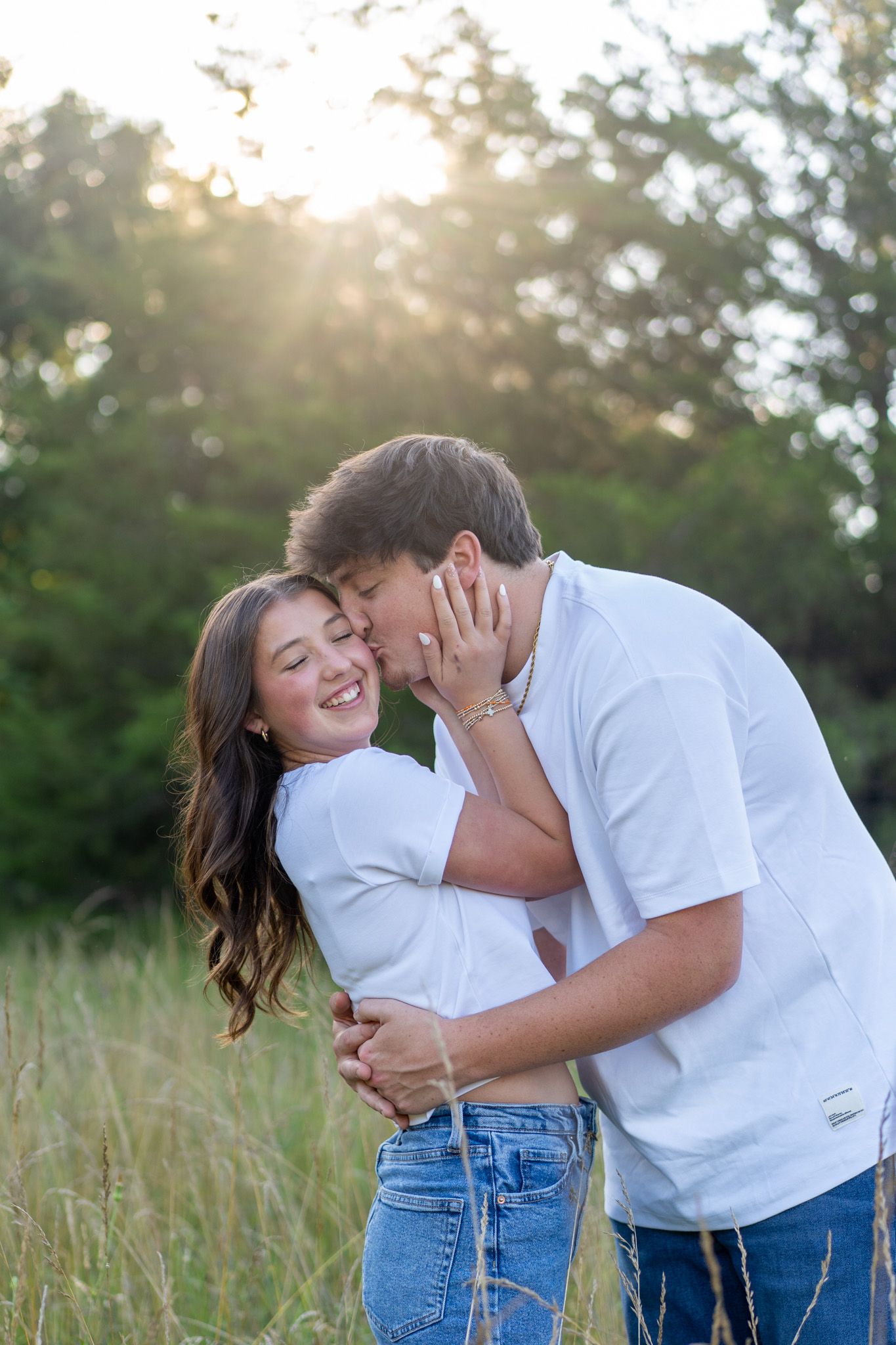 A young man in a white shirt kisses a laughing girl on the cheek in a sun-drenched grassy field.