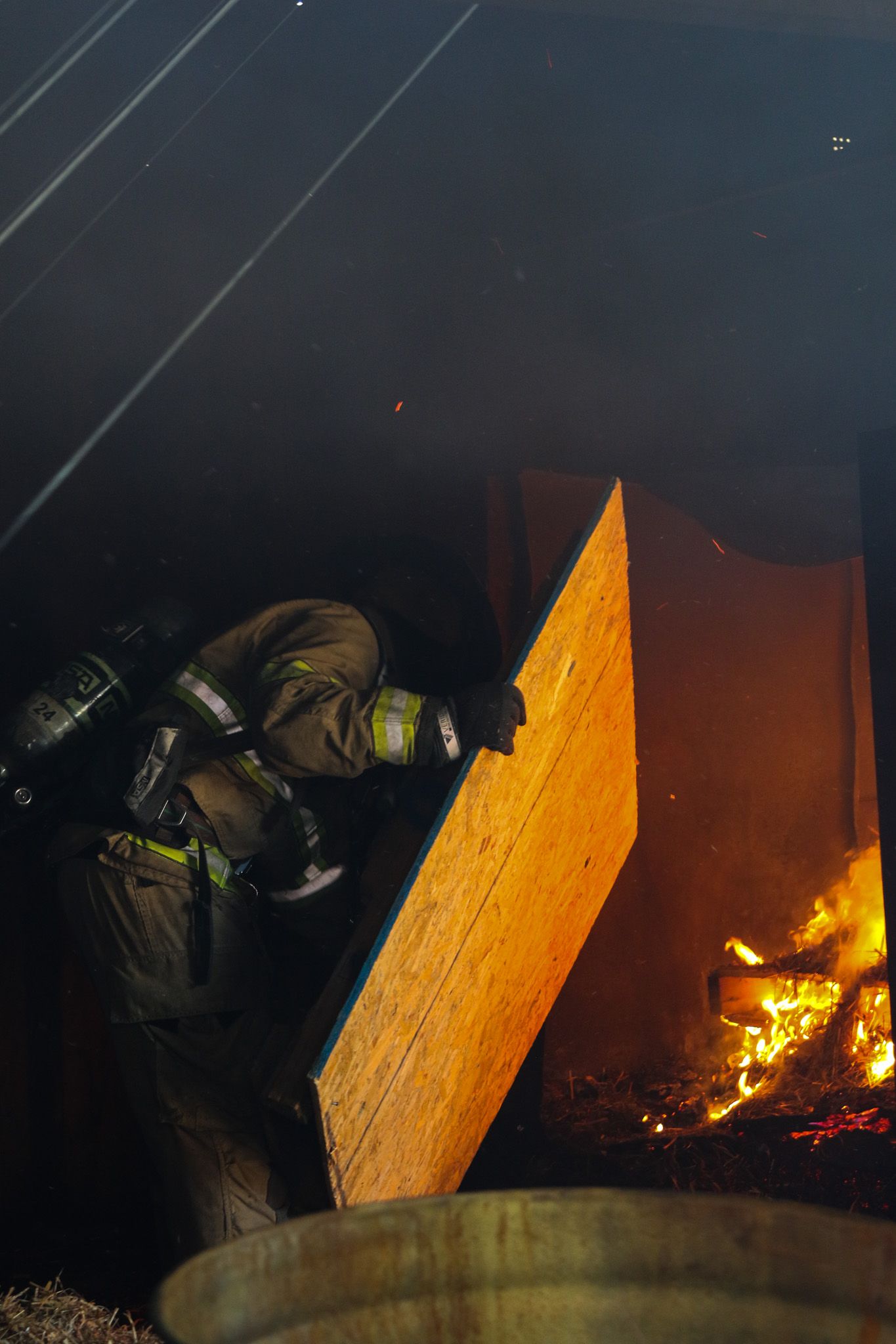 Firefighter in full gear and breathing apparatus battles a structural fire, lifting plywood amid flames and thick smoke.