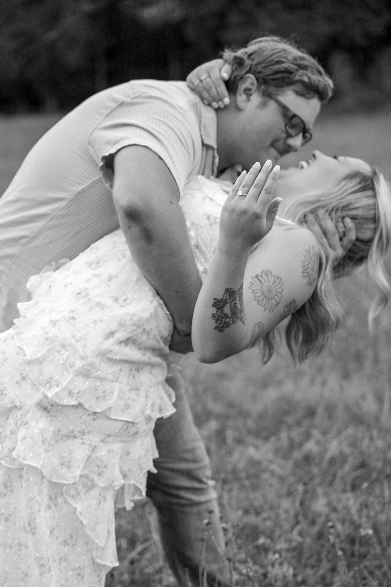 Black and white photo of man dipping and kissing tattooed blonde woman in ruffled dress in field.