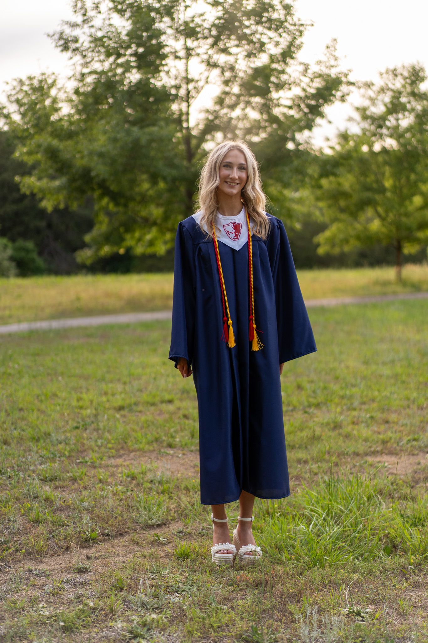 Young woman in navy blue graduation gown with gold tassels smiling outdoors in a green park setting.