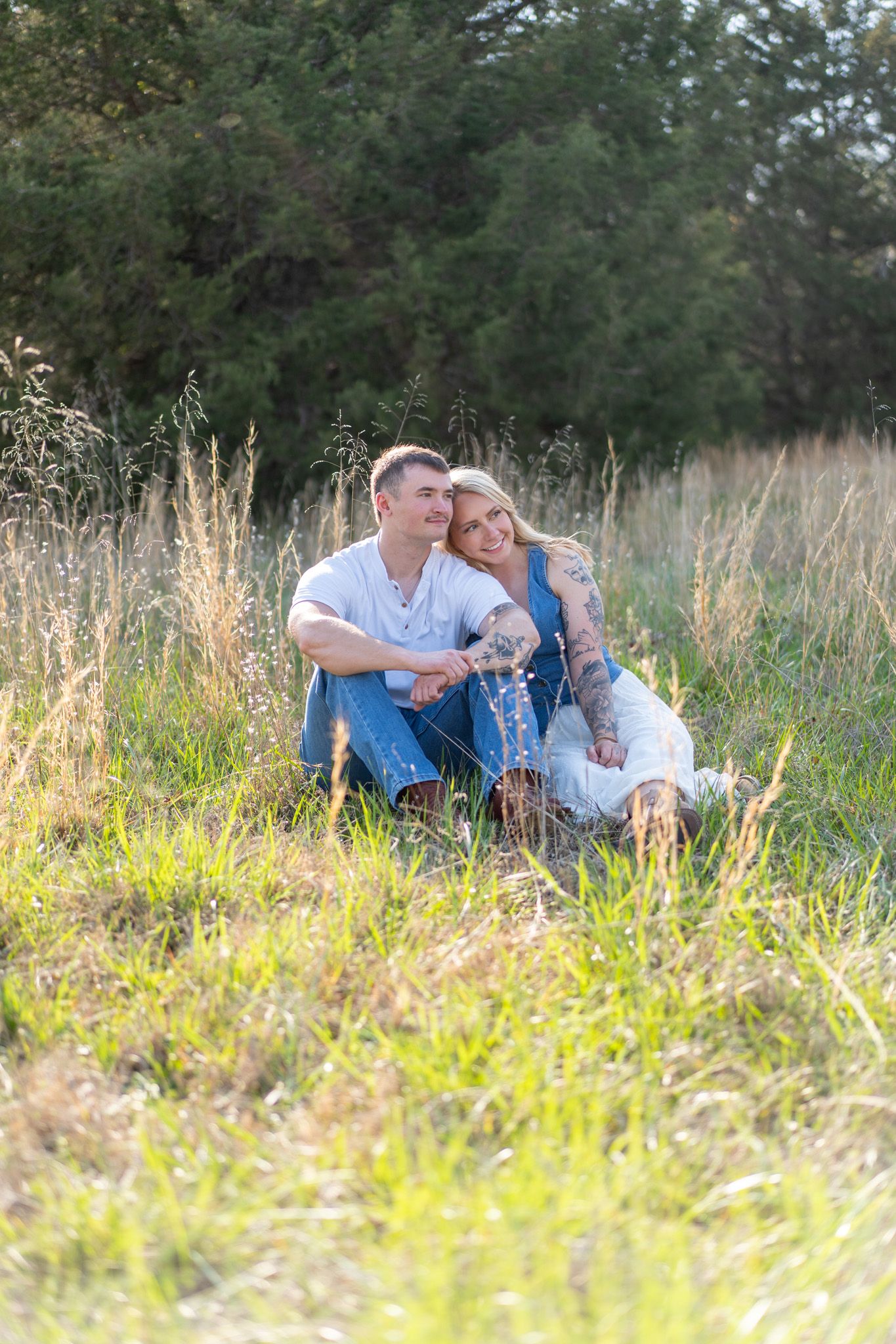 Happy couple sitting together in sunny golden meadow with tall grass and trees.