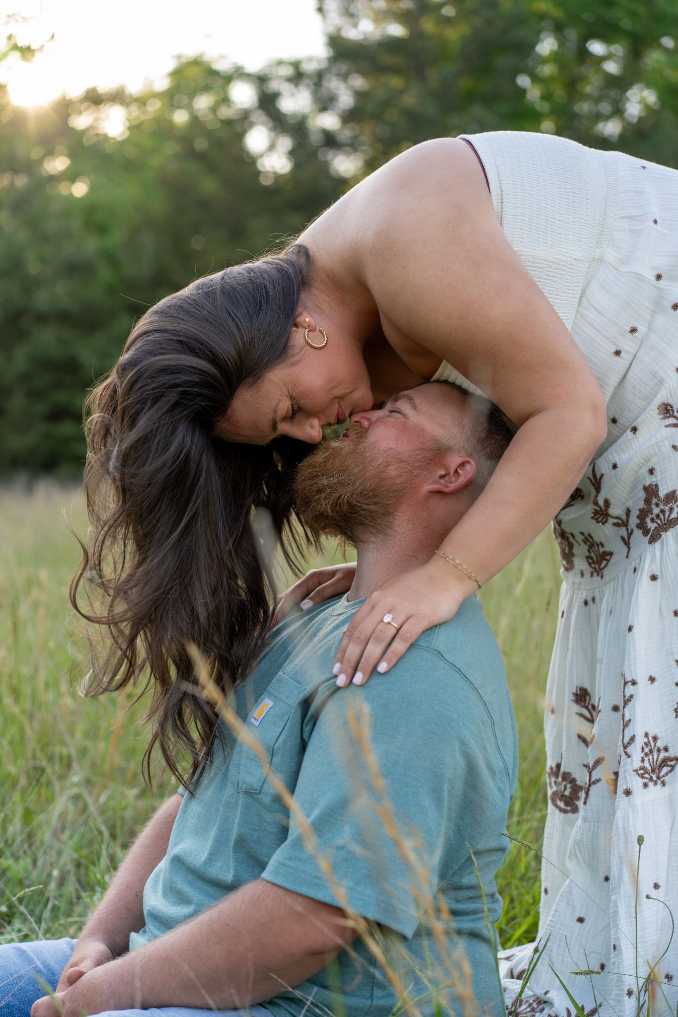 Engaged couple sharing a romantic kiss in a sunlit grassy meadow.