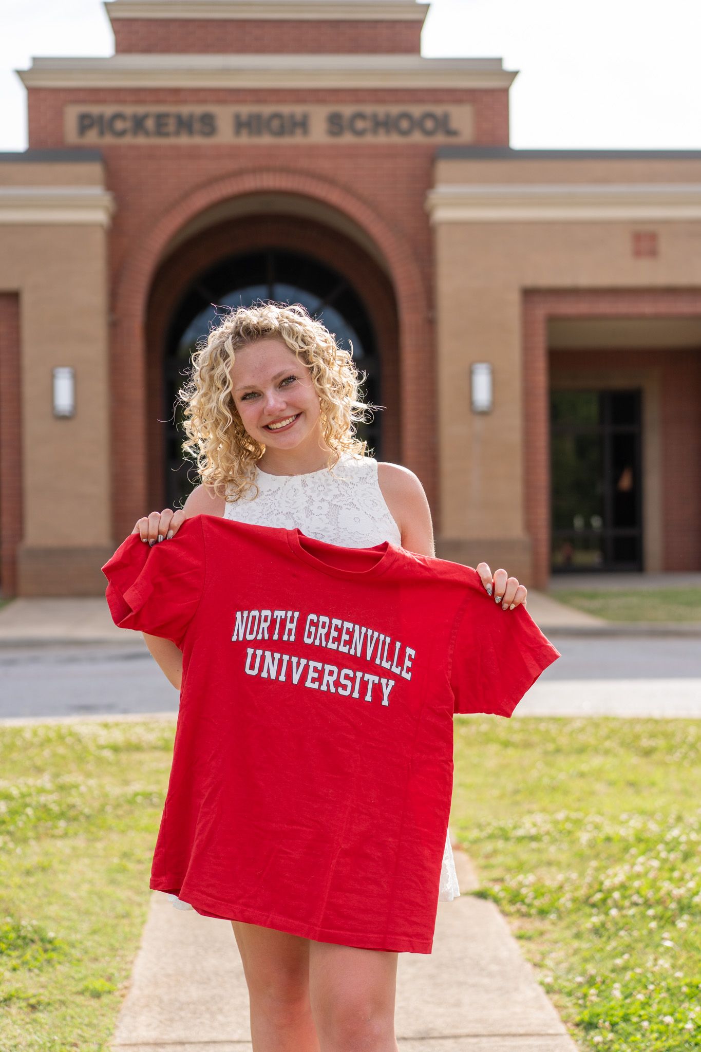 Smiling blonde girl holds North Greenville University red shirt in front of Pickens High School entrance.