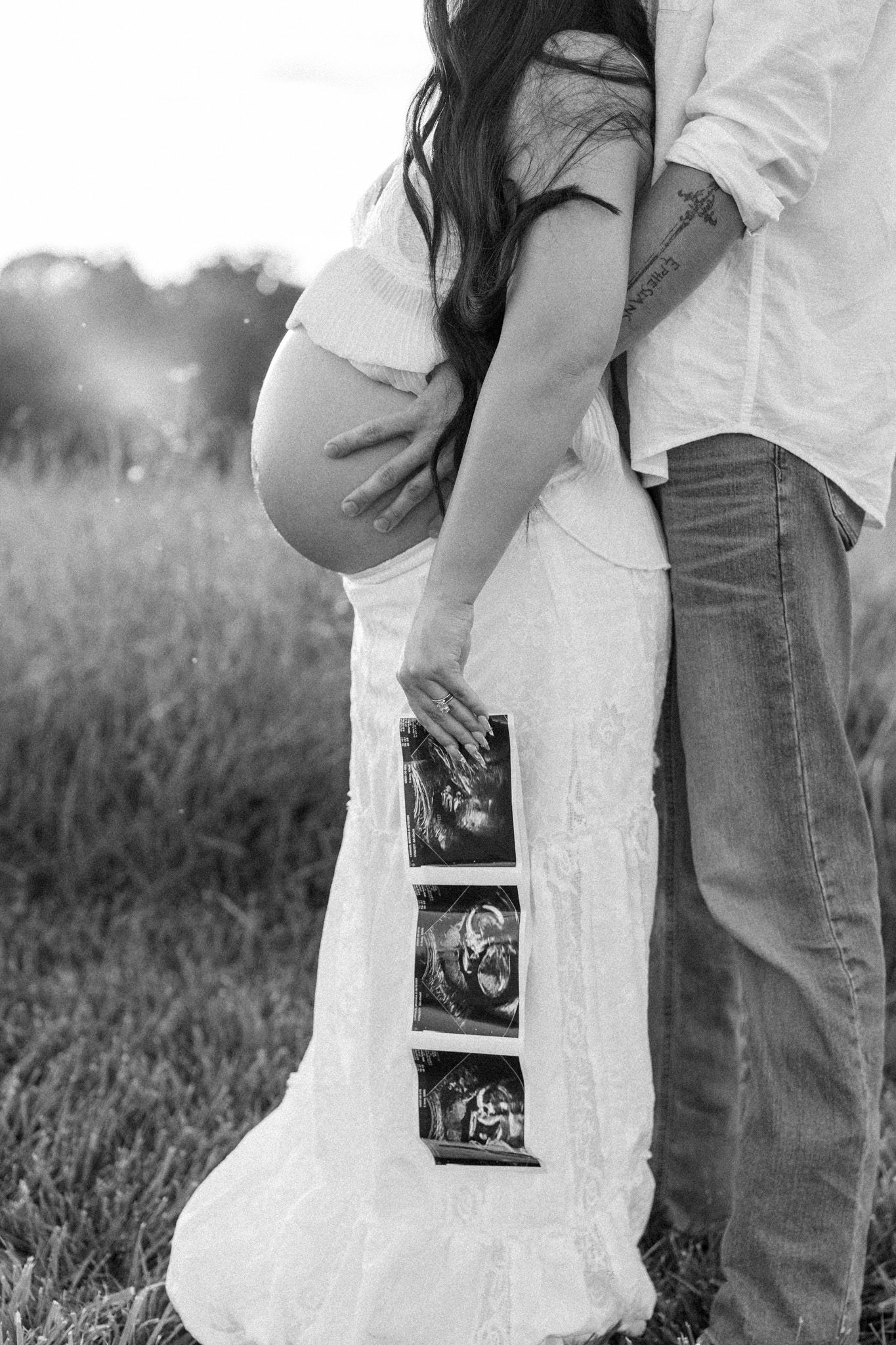 Black and white maternity photo of couple holding ultrasound images in a grassy field.
