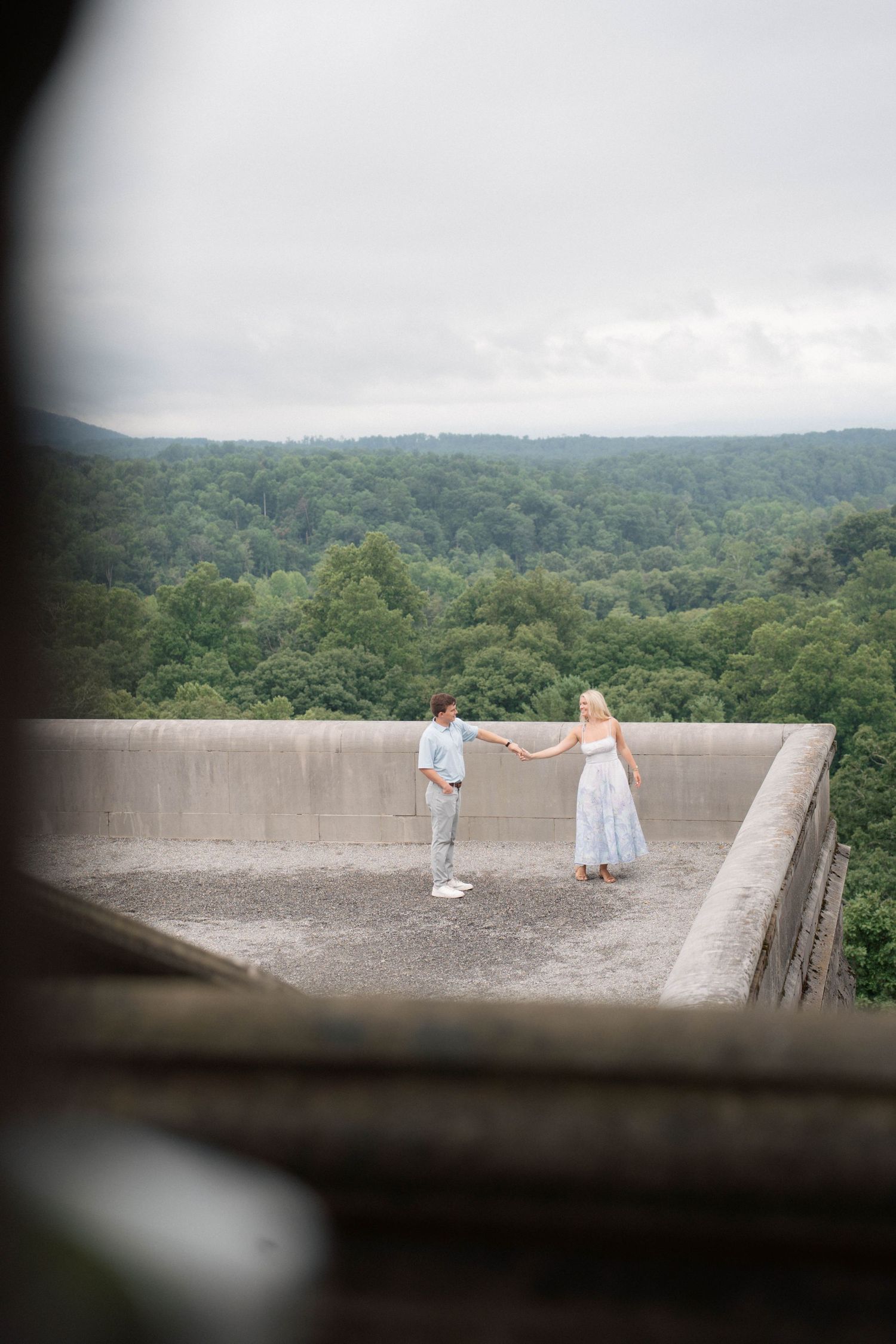 Engaged couple holding hands on scenic concrete overlook surrounded by lush green forest.