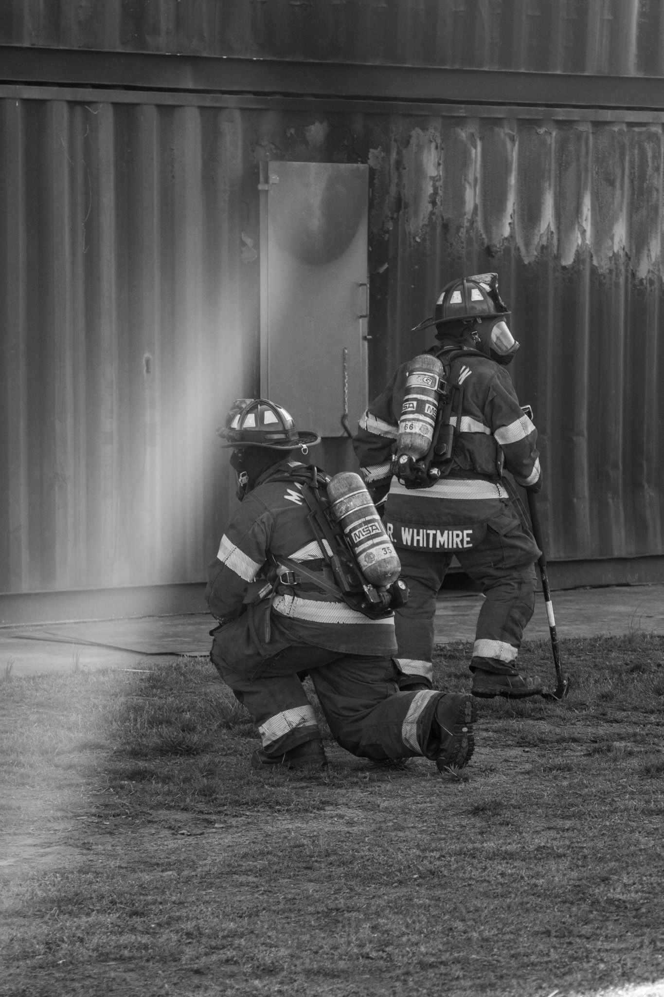 Two firefighters in full gear crouch near a metal building door during a training exercise, one named Whitmire visible on their jacket.