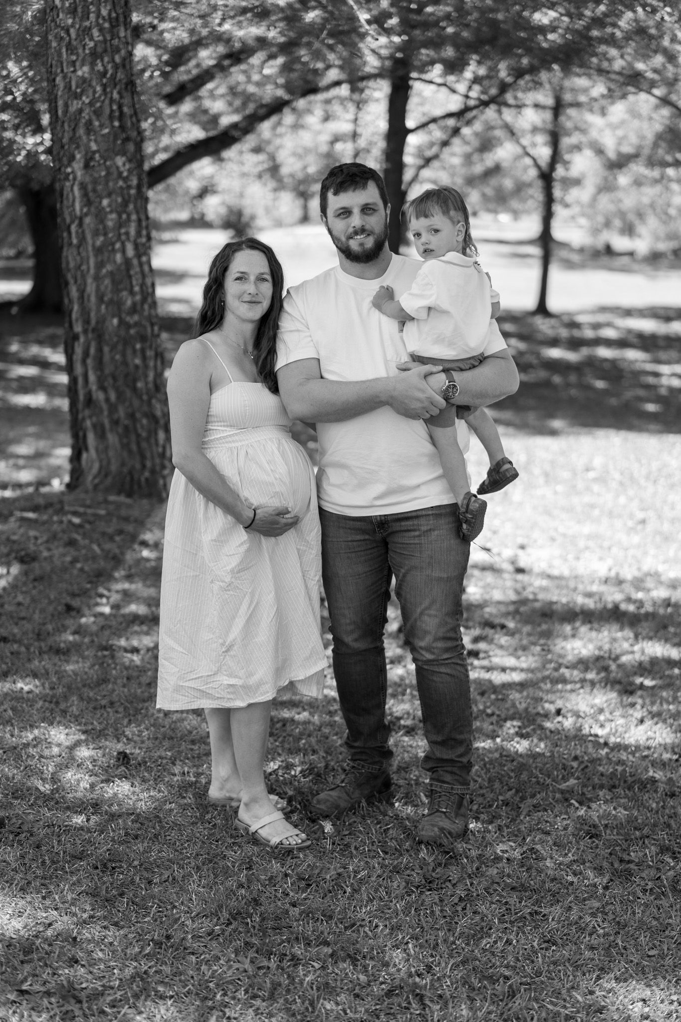 Black and white photo of a pregnant woman and man holding a toddler posing together outdoors in a park.