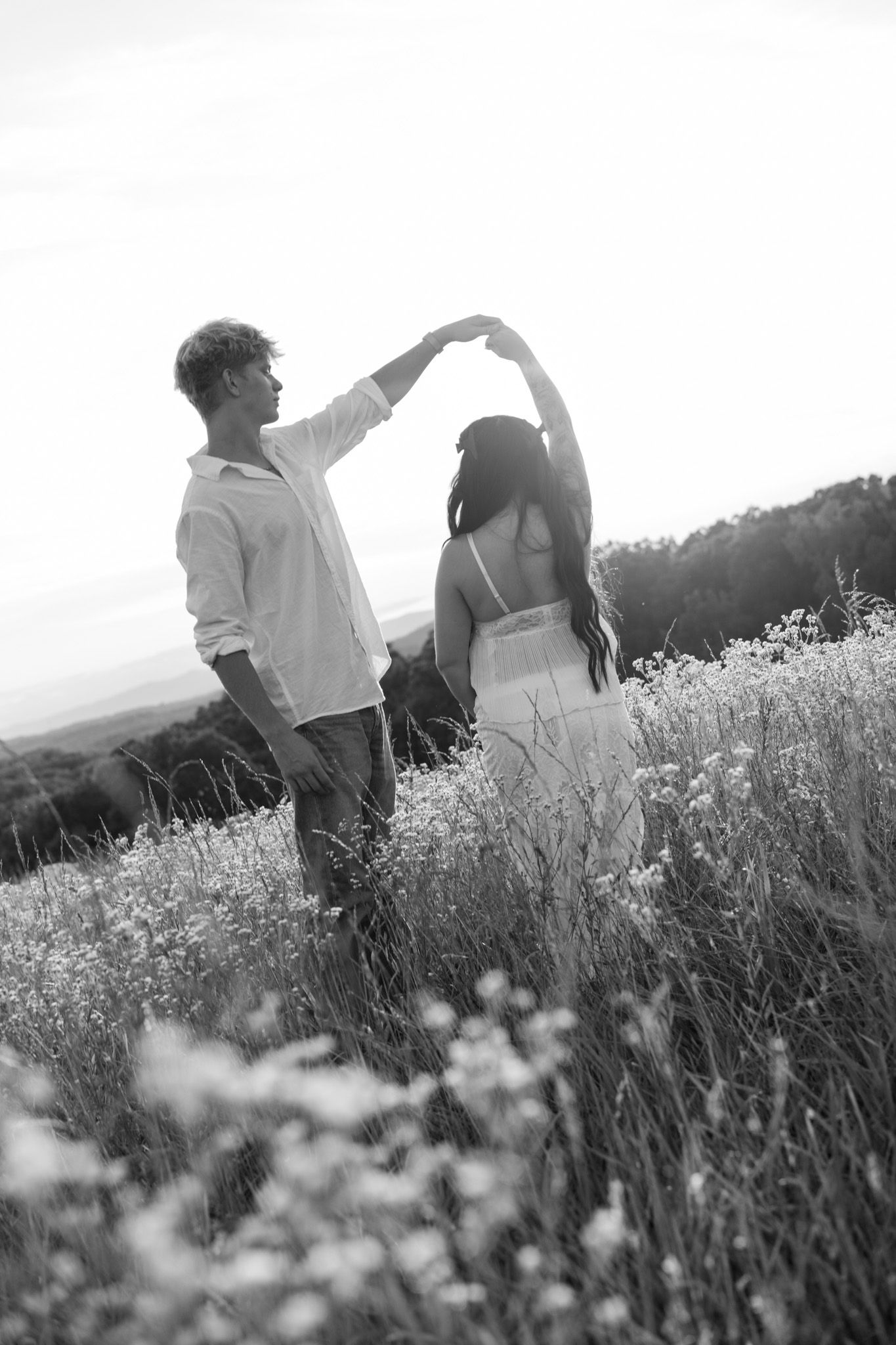 Black and white photo of couple dancing in wildflower field, man spinning pregnant woman at sunset.