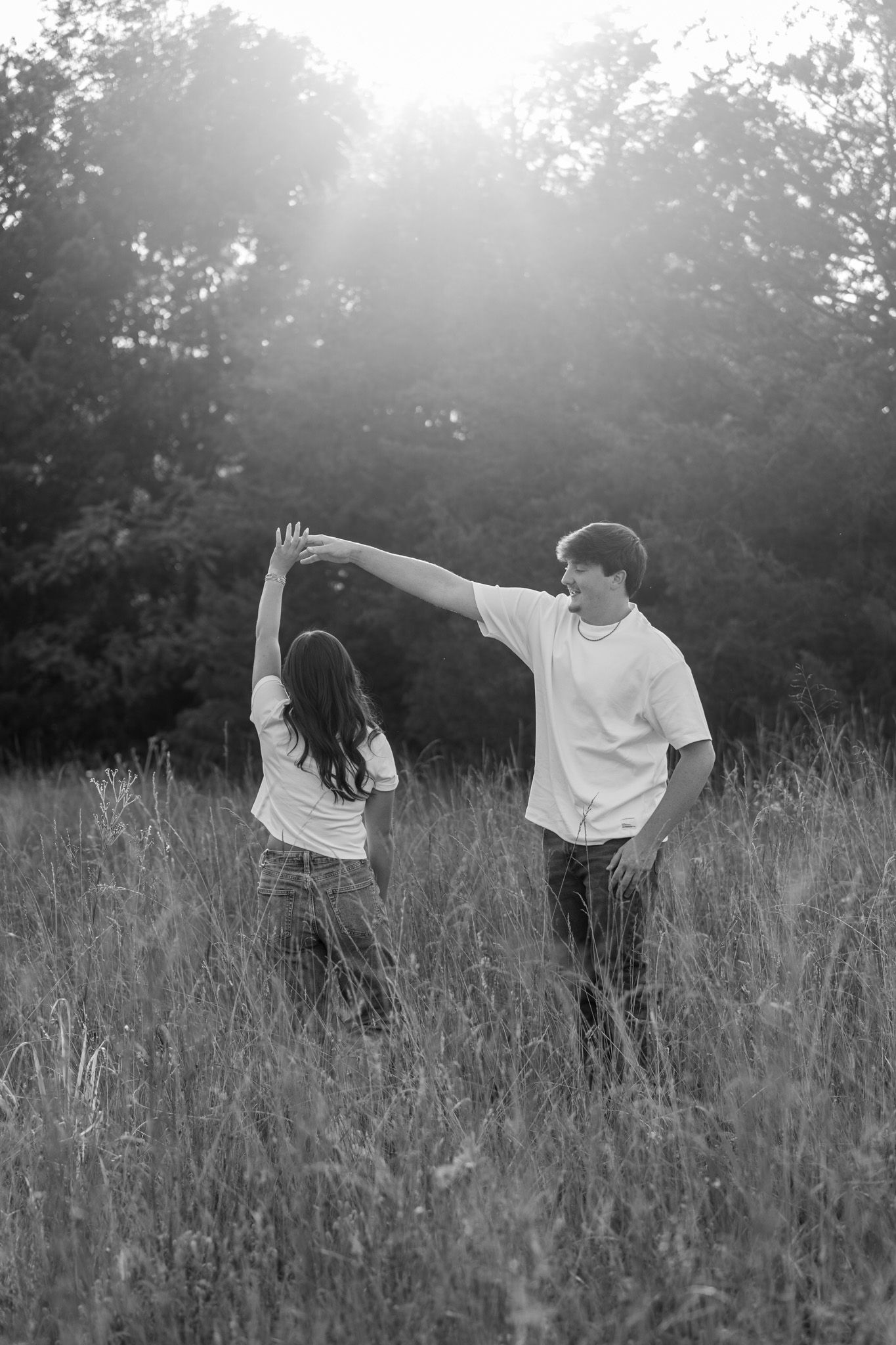 Black and white photo of a young couple dancing playfully in a tall grass field with glowing backlight.