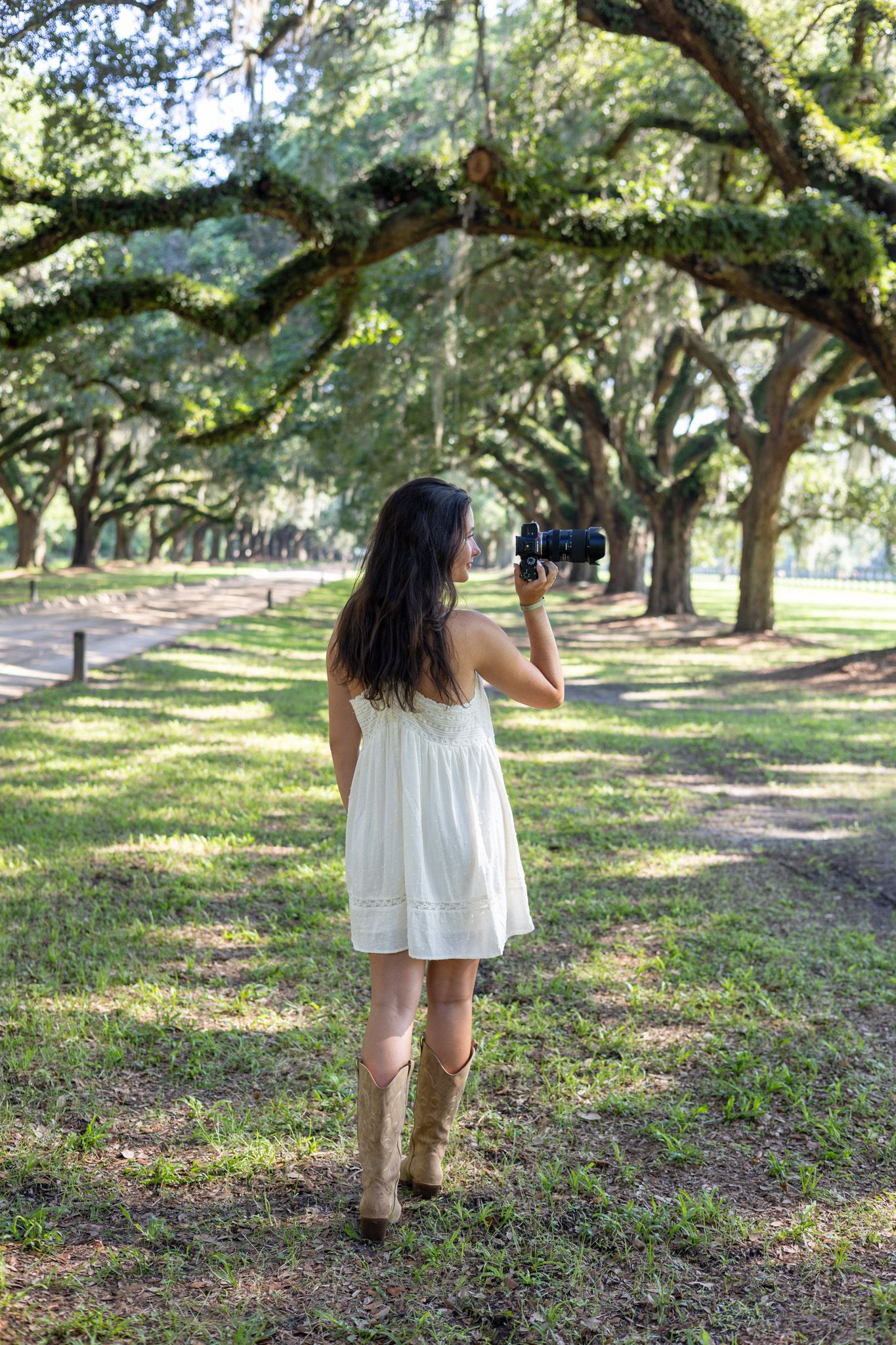 Woman in white dress and cowboy boots photographing moss-covered oak tree alley in a lush green park.