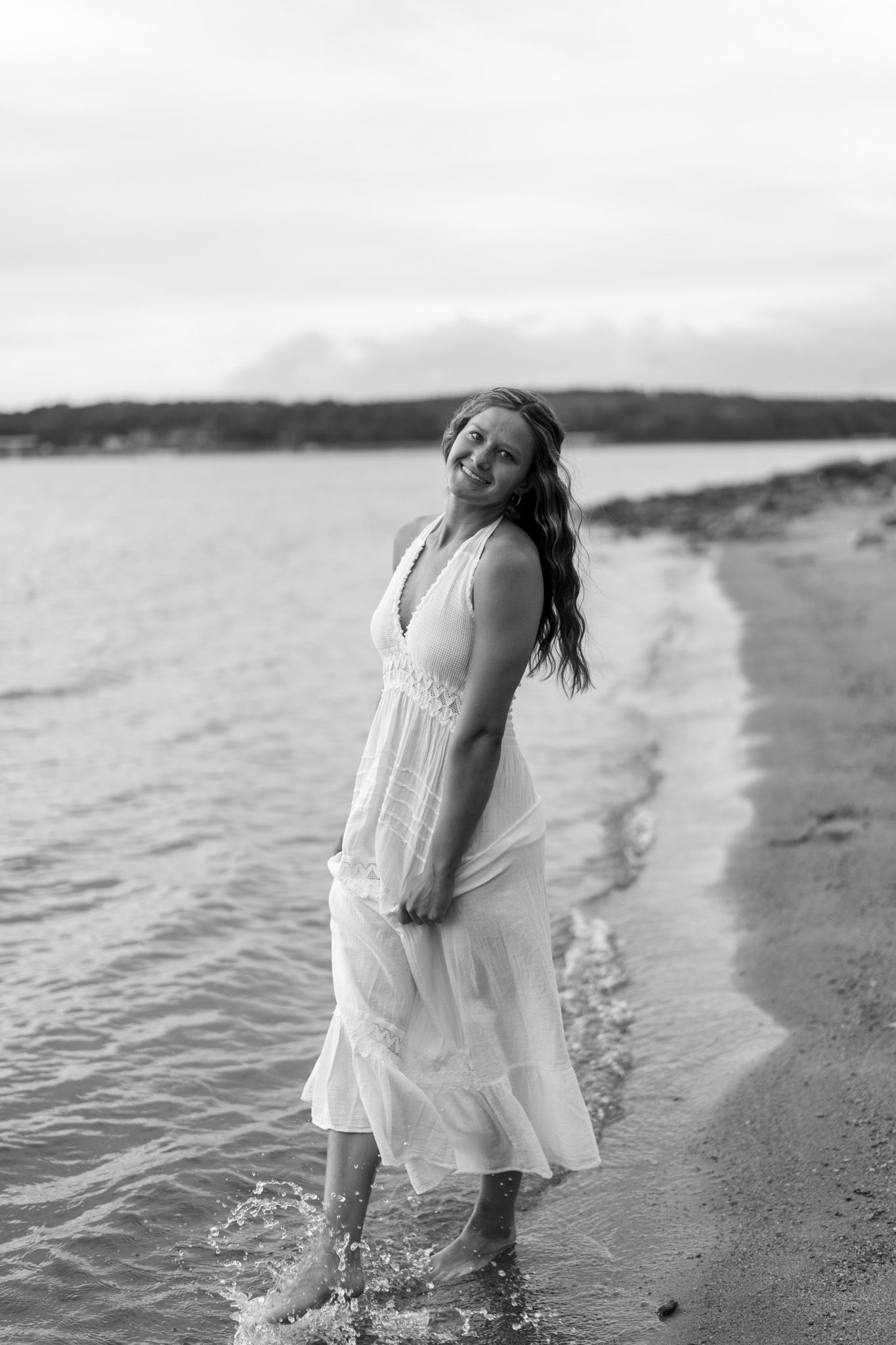 Young woman in white dress walking along a sandy beach shoreline in black and white photo.