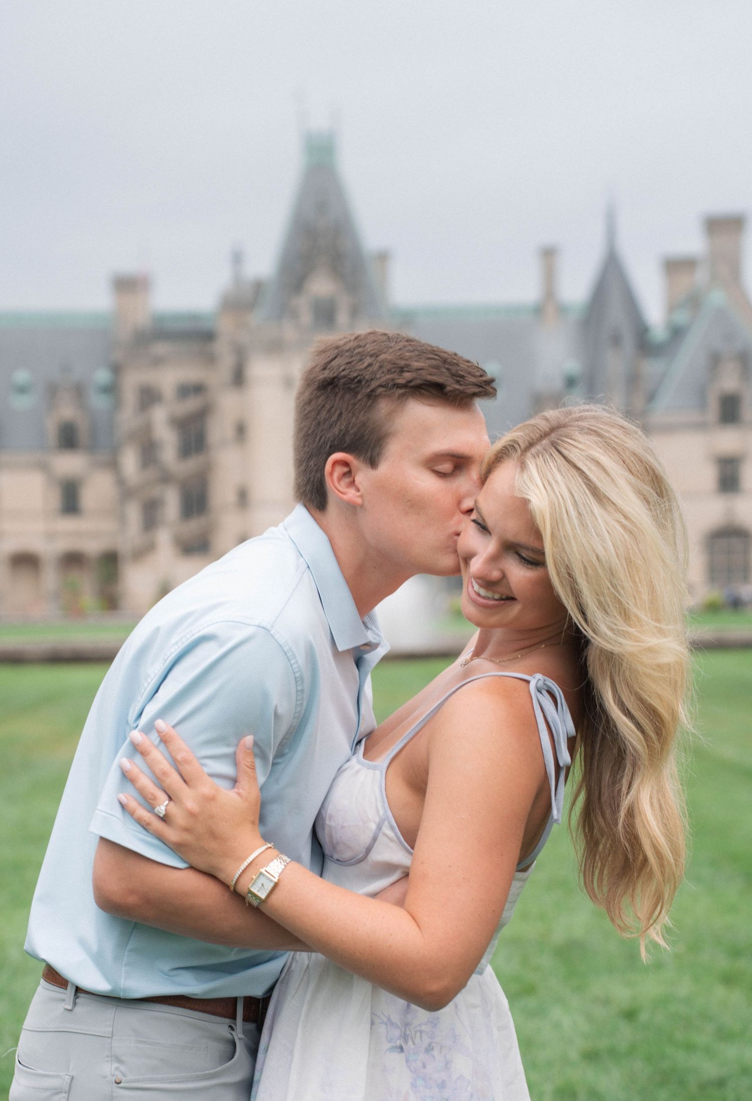 Man kissing smiling blonde woman on cheek with grand Biltmore Estate castle visible behind them.