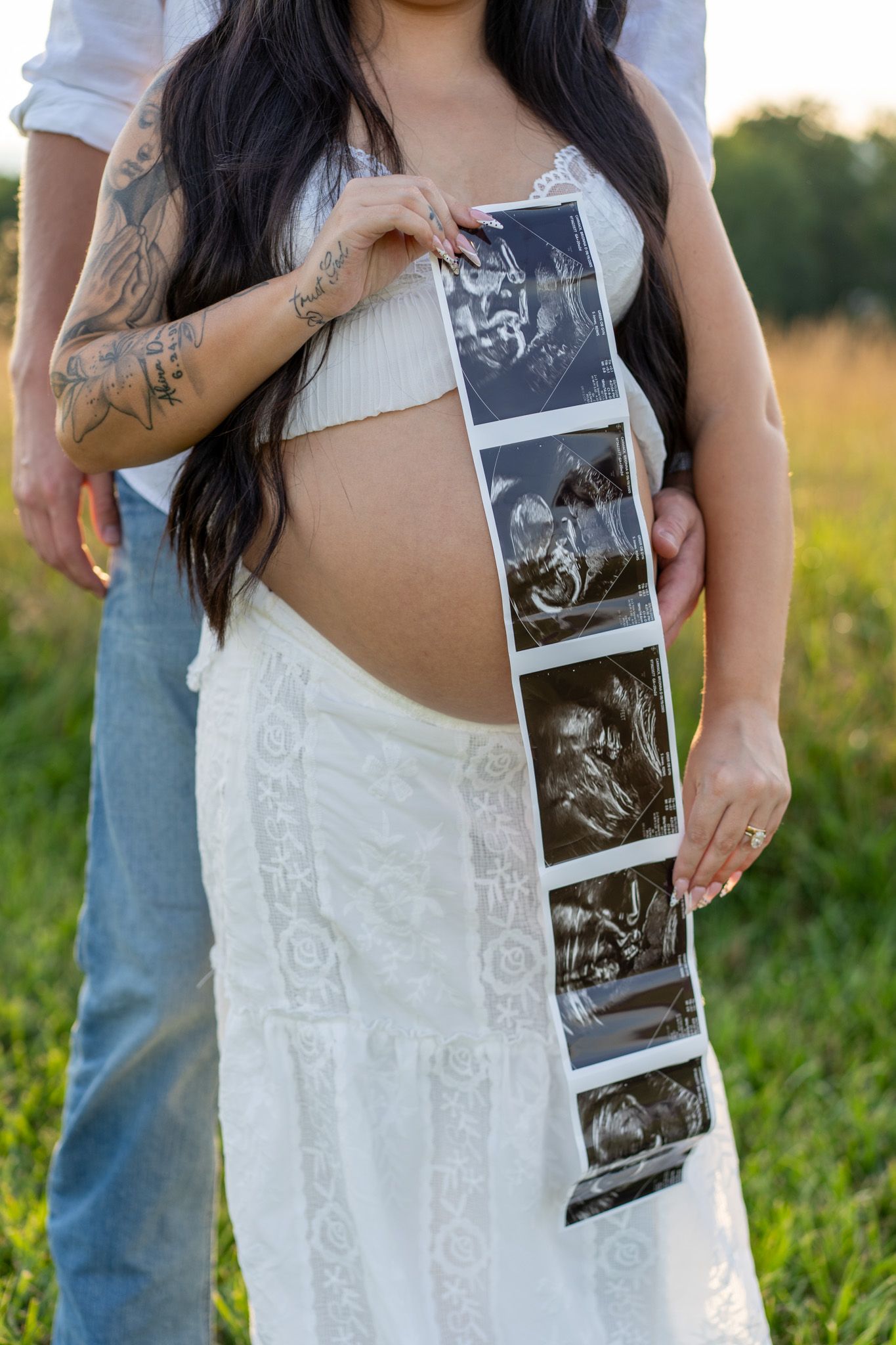 Pregnant woman in white lace dress holding ultrasound strip photos against her bare baby bump in a field.