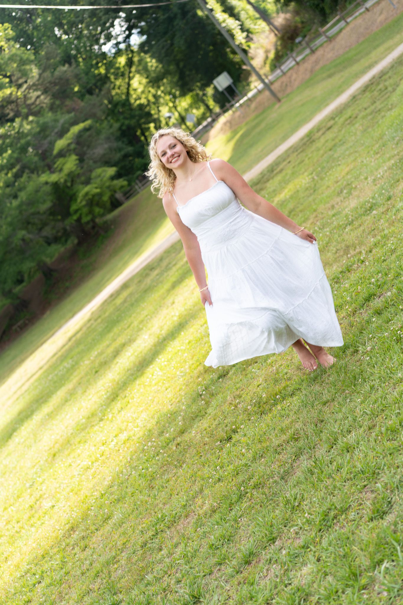 Smiling blonde woman in a flowing white dress walking barefoot on green grass in a sunny outdoor setting.