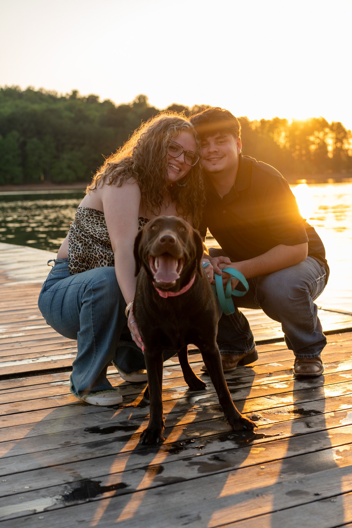 Smiling couple crouching with chocolate Labrador on wooden dock at sunset.