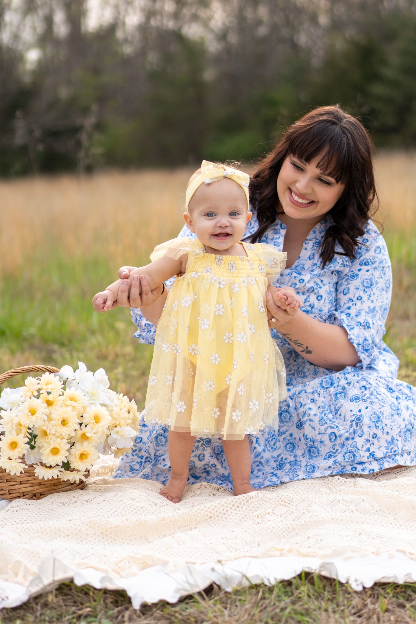 Smiling mother in blue dress supports standing baby girl in yellow floral dress on outdoor picnic blanket.