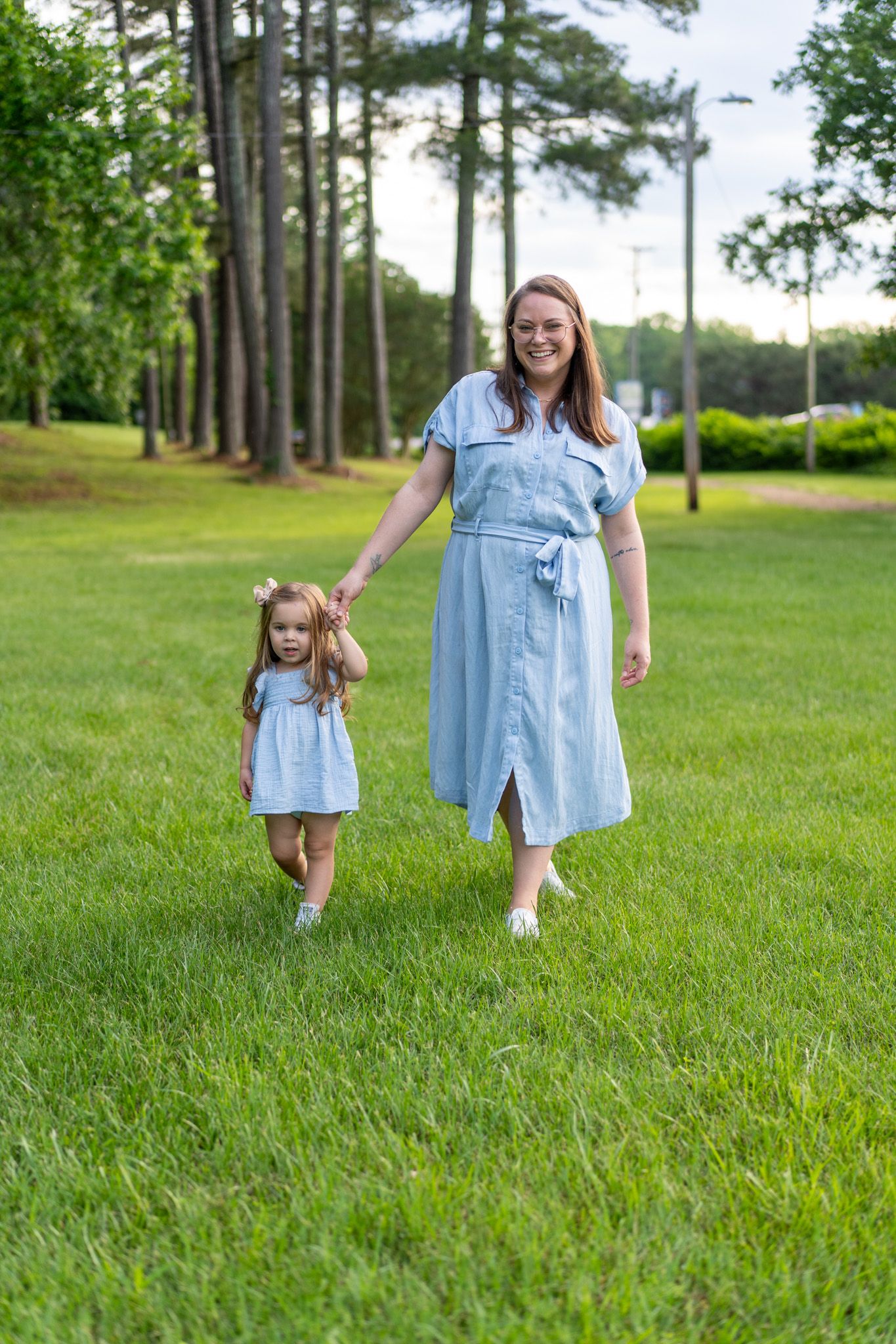 Smiling mother in blue dress walking hand-in-hand with toddler daughter in green park.