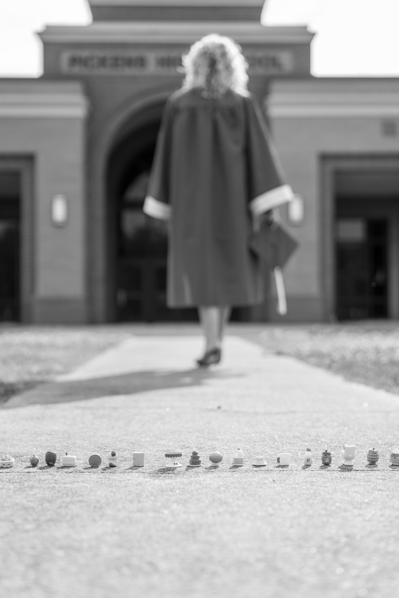 Black and white photo of graduate in cap and gown walking toward school entrance with pebbles in foreground.