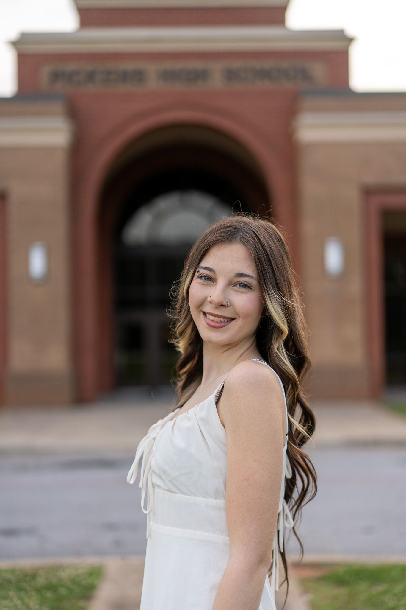 Smiling young woman in white dress poses in front of McKenzie High School brick entrance for senior photos.