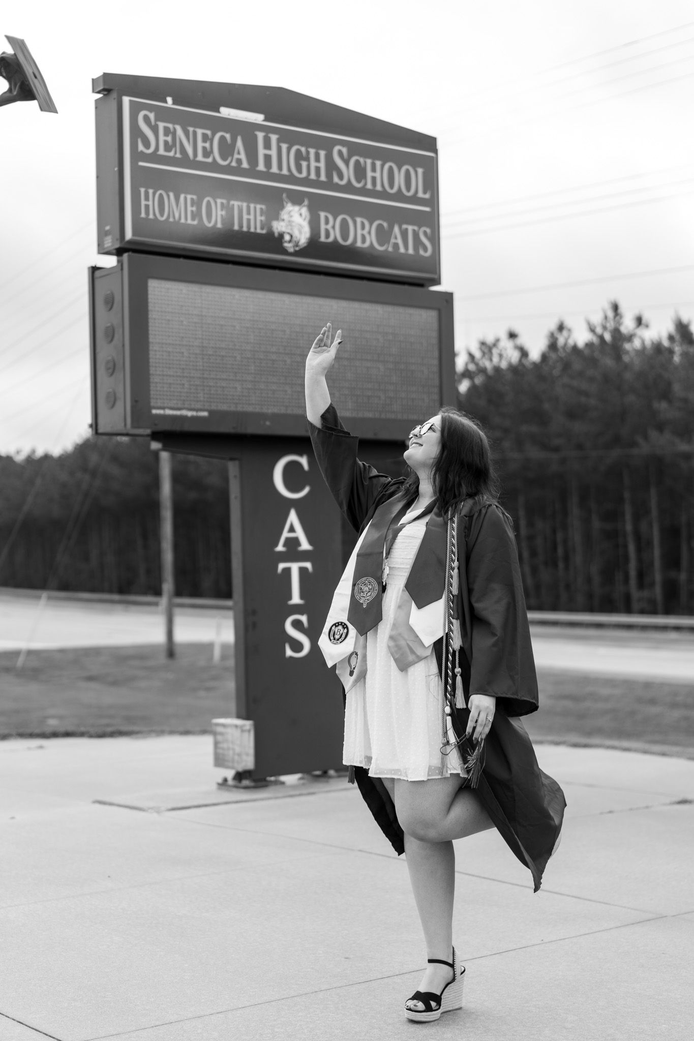 Black and white photo of graduate posing by Seneca High School Bobcats sign in cap and gown.