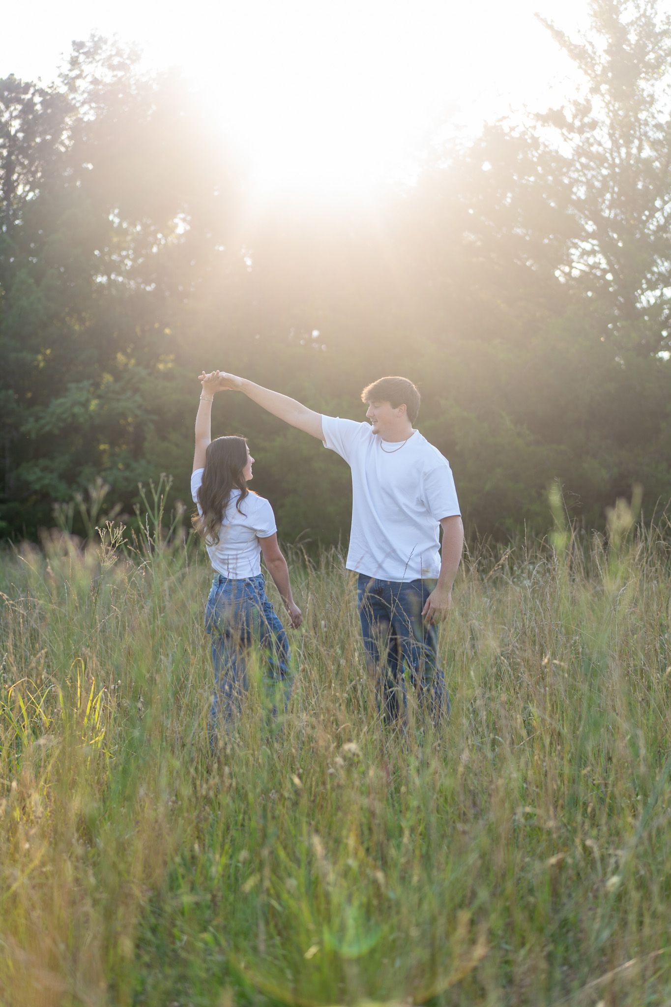 Couple dancing in sunlit meadow, man spinning woman in matching white shirts and jeans at golden hour.