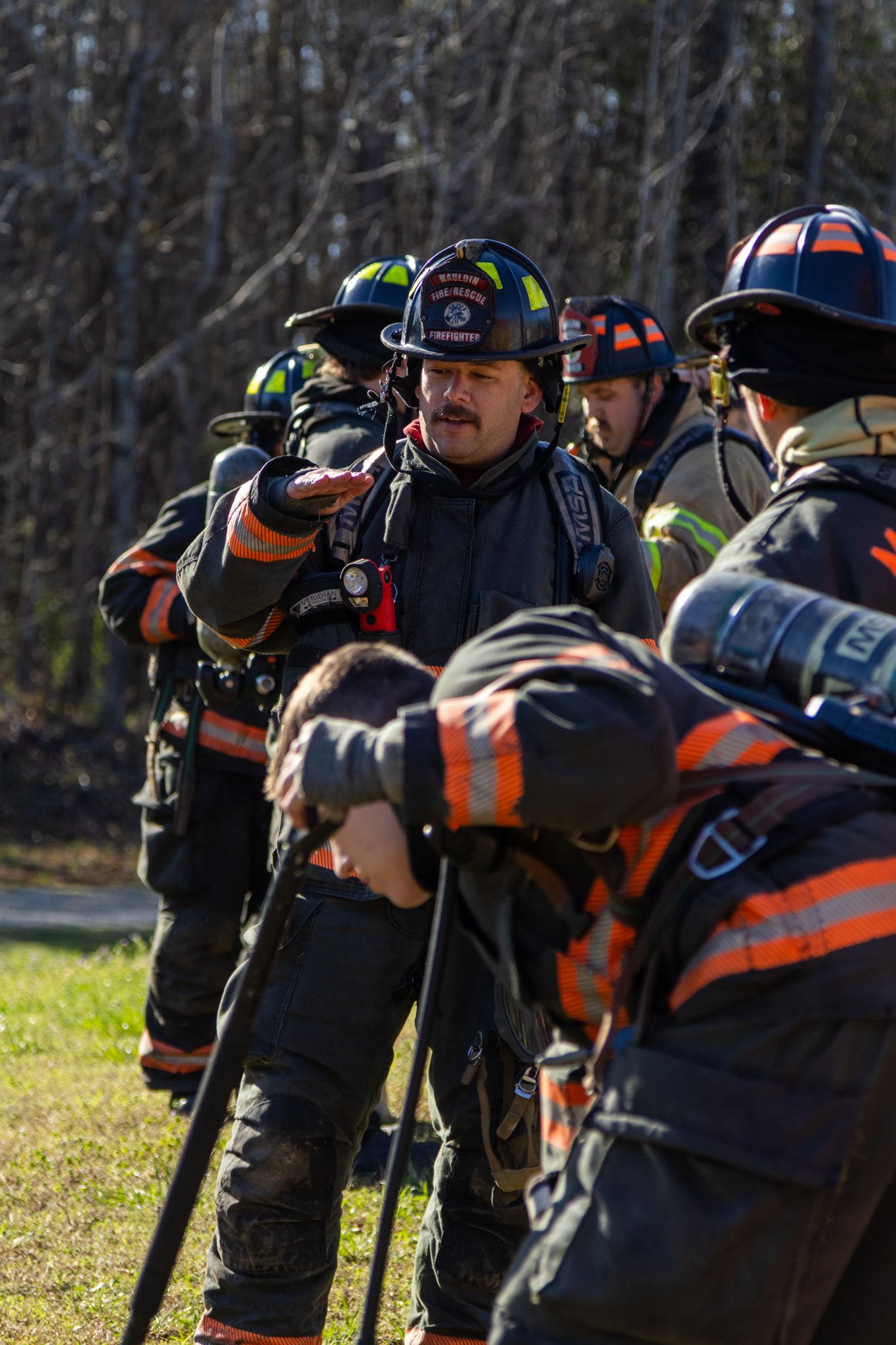 Group of firefighters in black and orange turnout gear during an outdoor training exercise.