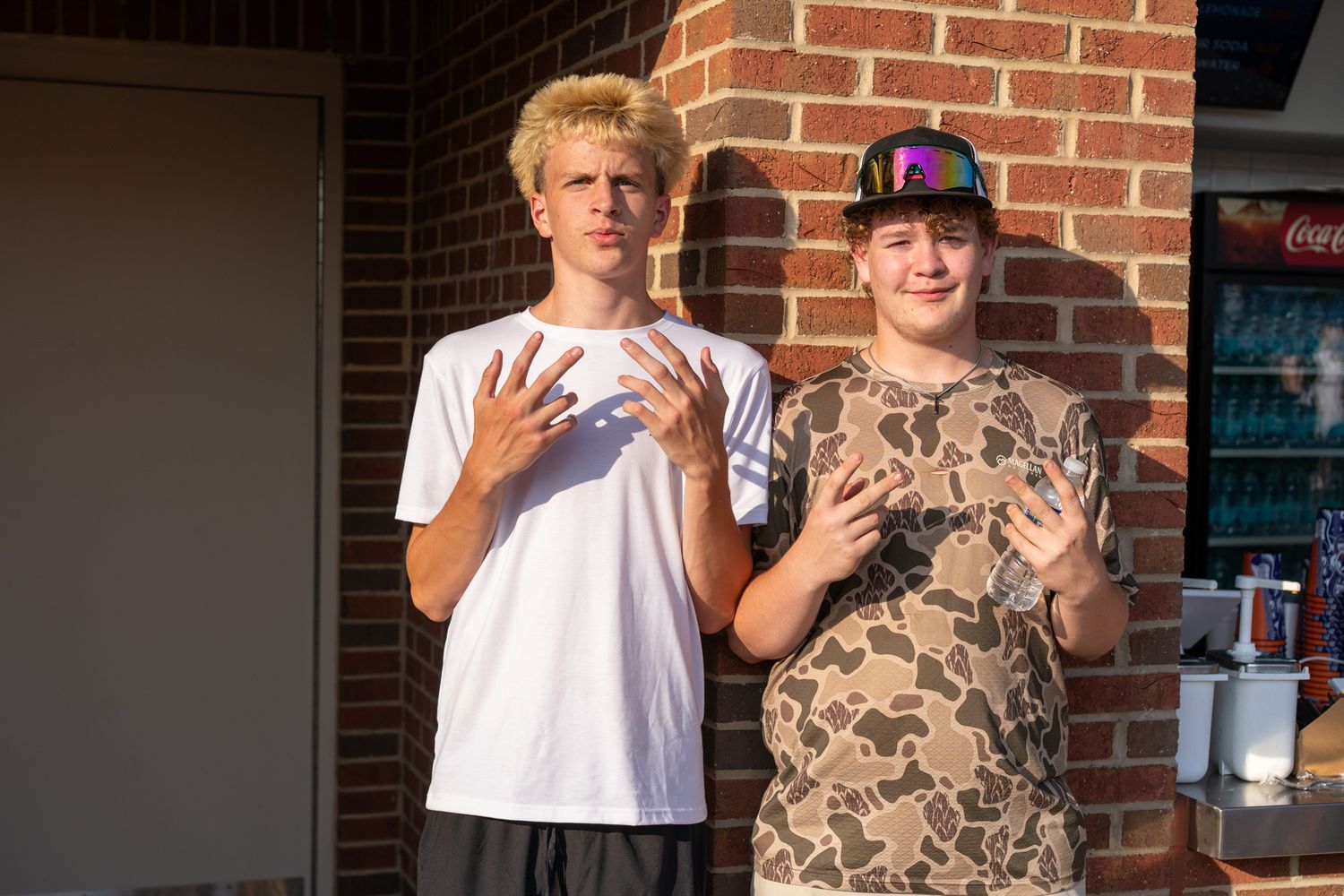 Two teenage boys posing outdoors near a brick wall, making hand gestures, one in white tee and one in camo shirt.
