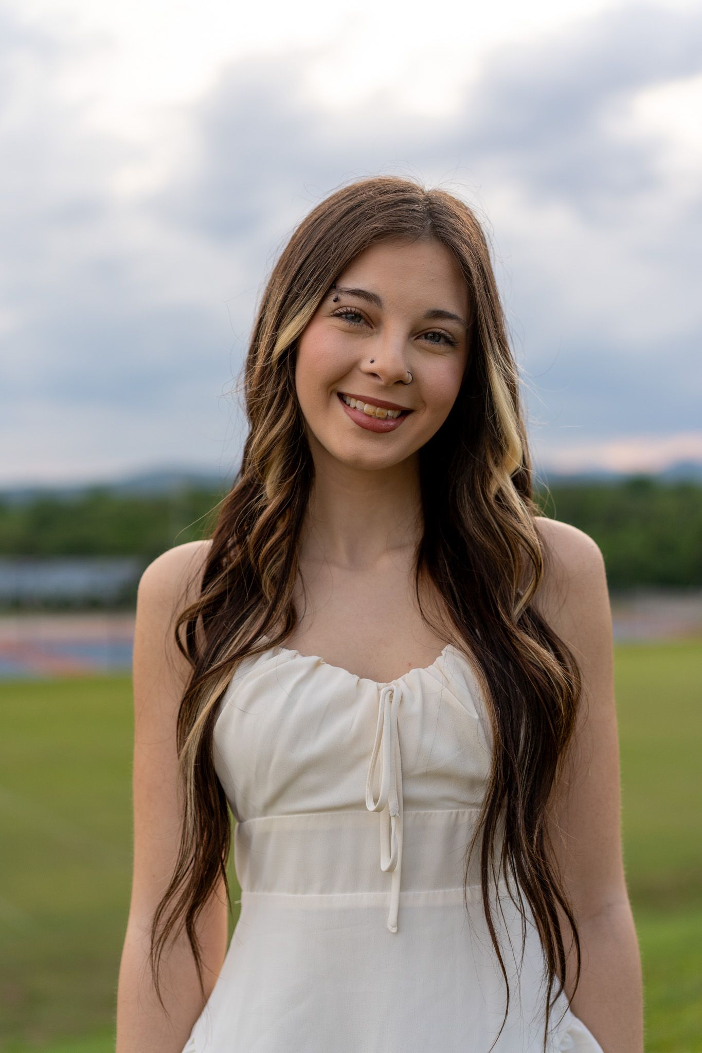 Young woman with long wavy brown hair smiling outdoors in a white sundress against a cloudy sky.