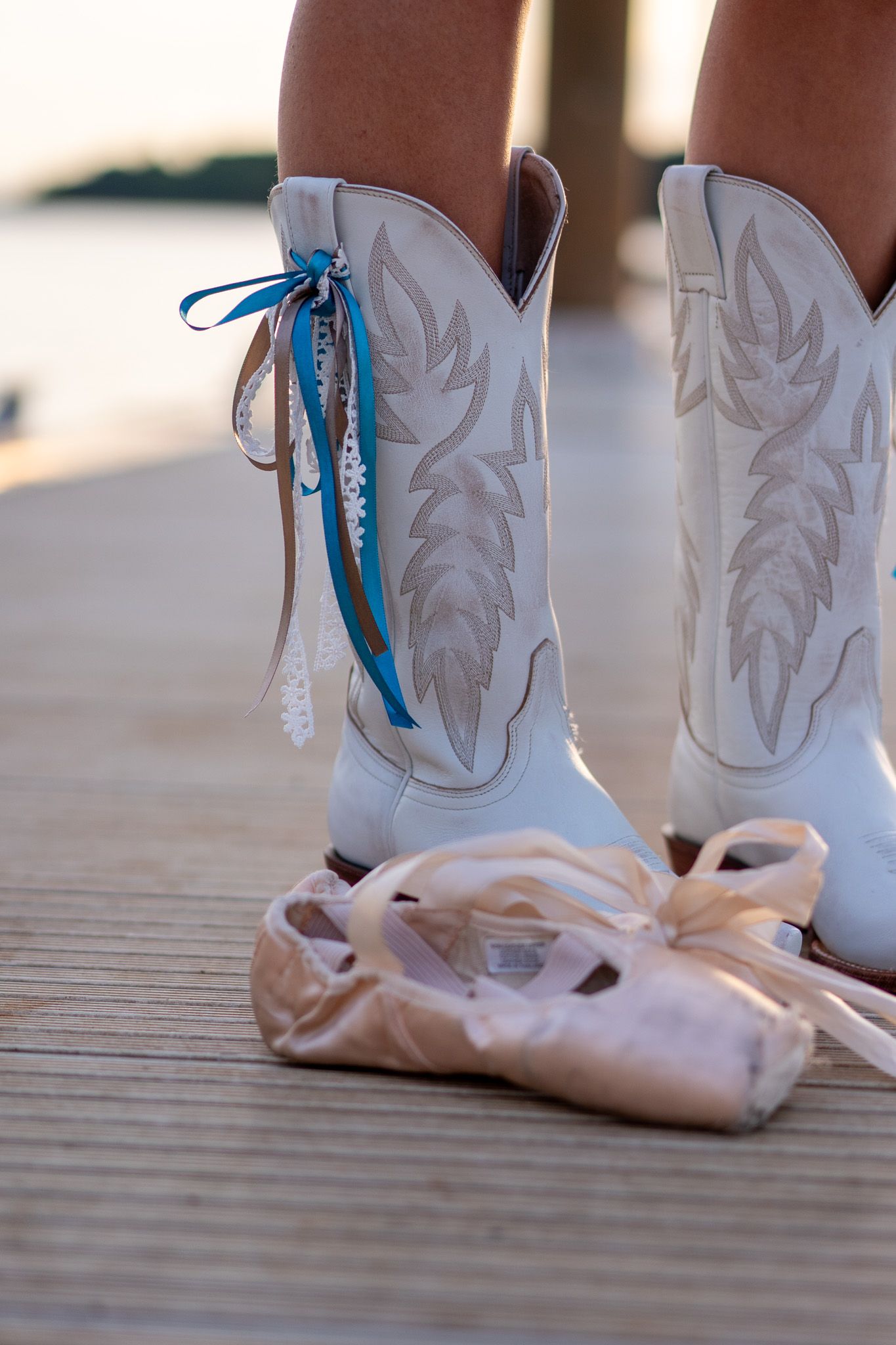 White cowboy boots with blue ribbon beside ballet pointe shoes on wooden dock at sunset.