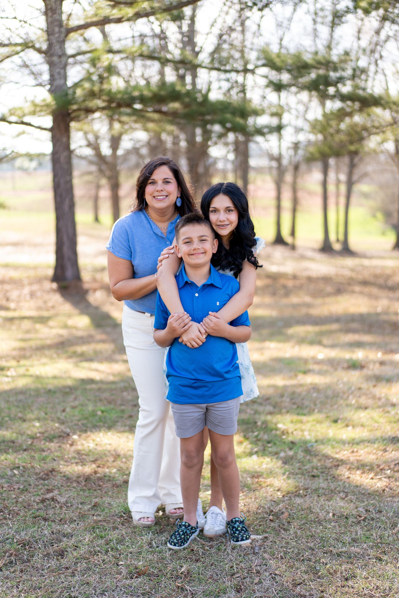 A smiling family of three poses outdoors among trees, with a woman in blue hugging a boy and teenage girl.