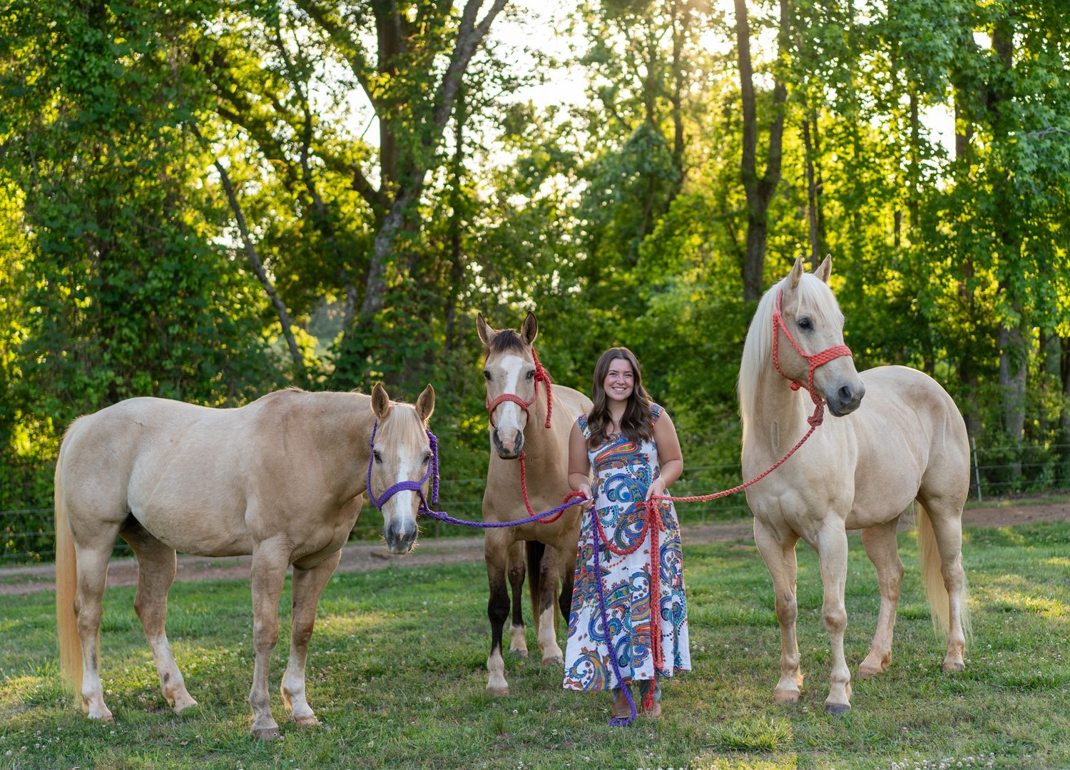 Smiling woman in floral dress standing with three palomino horses in sunny green pasture.