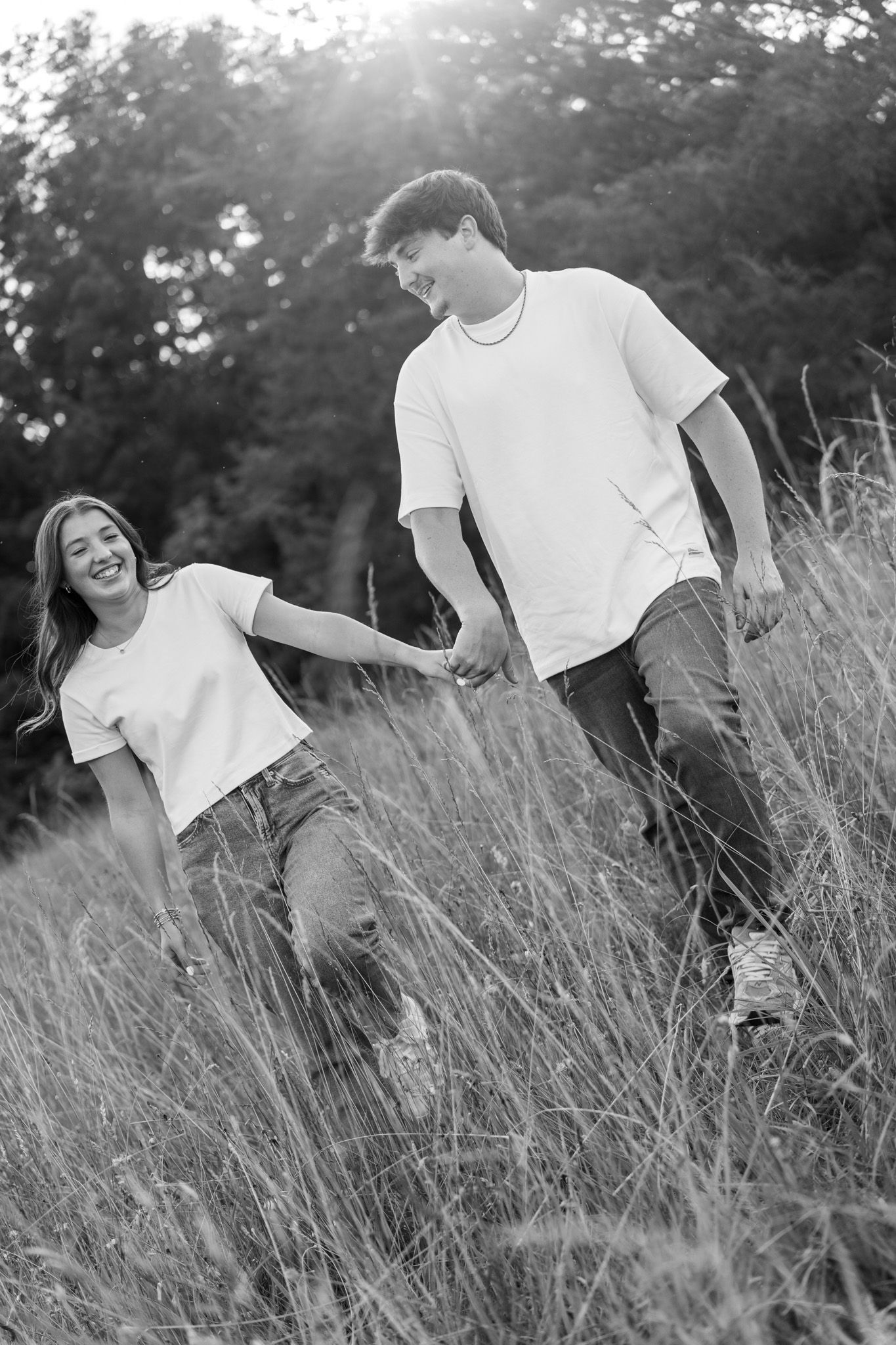 Black and white photo of young couple holding hands laughing while walking through tall grass field.