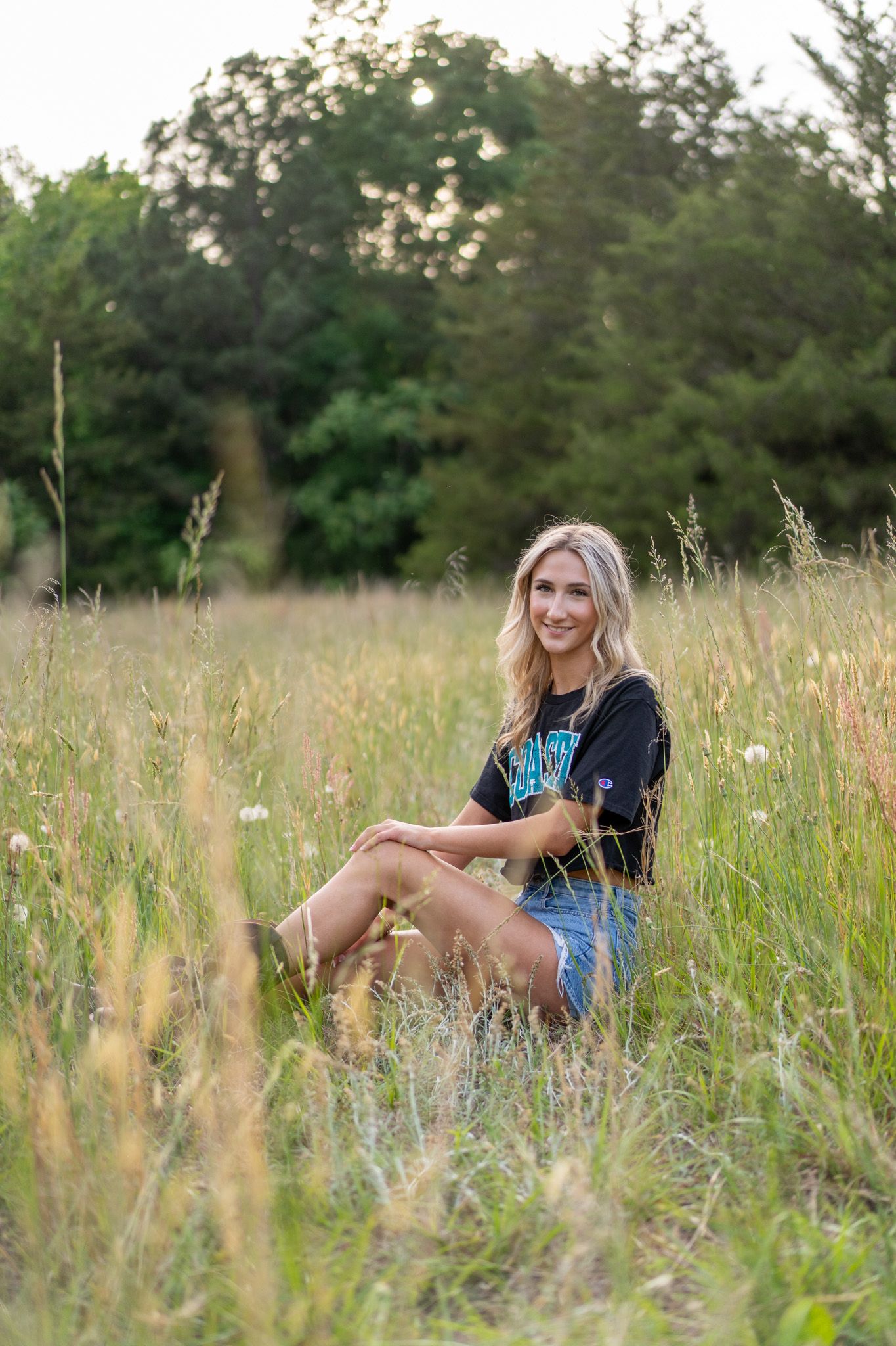 Young blonde woman sitting in tall grass field wearing denim shorts and dark graphic tee, smiling outdoors.