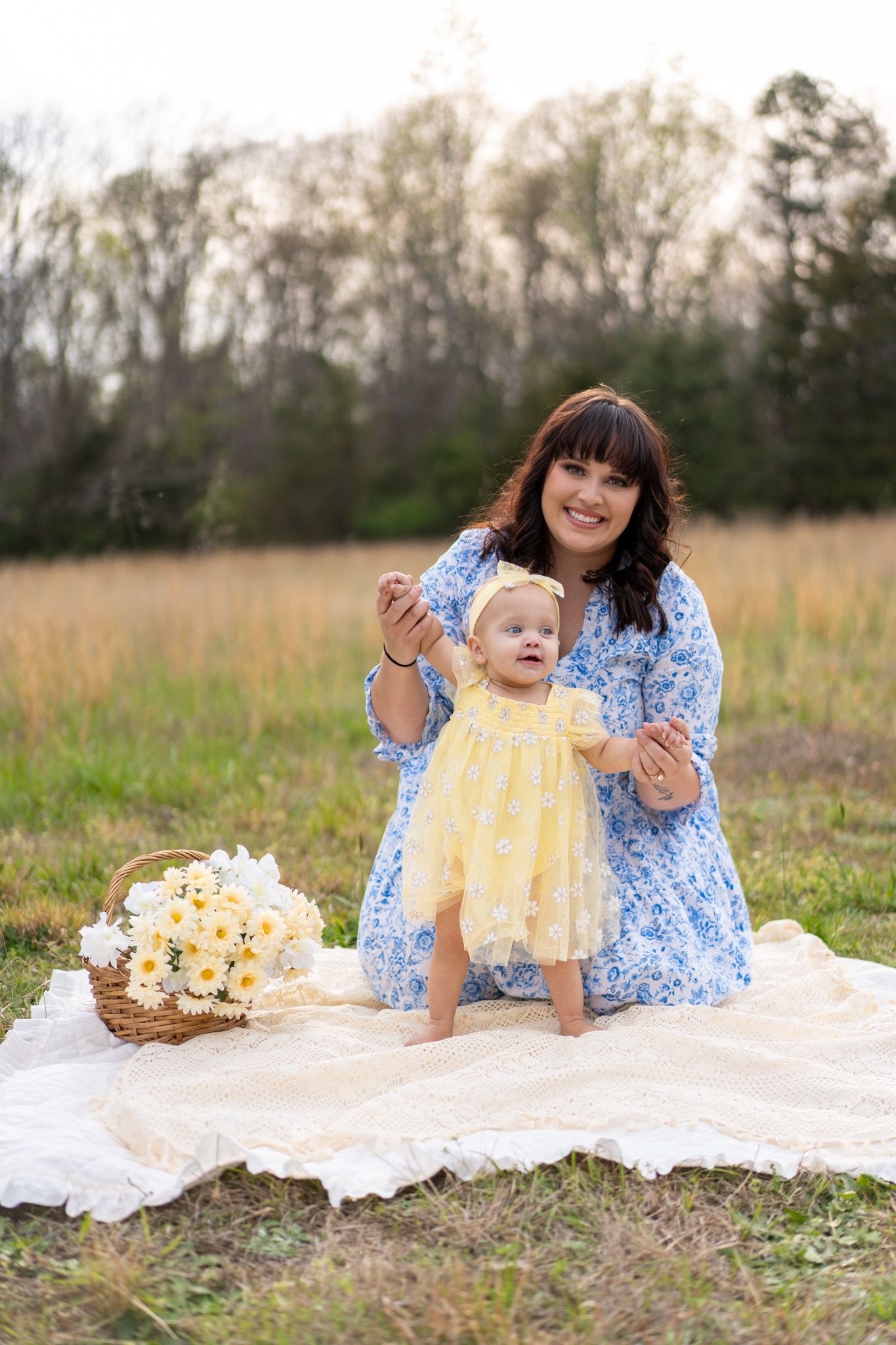Mother in blue floral dress helps baby girl in yellow dress stand on blanket in field with daisy basket.